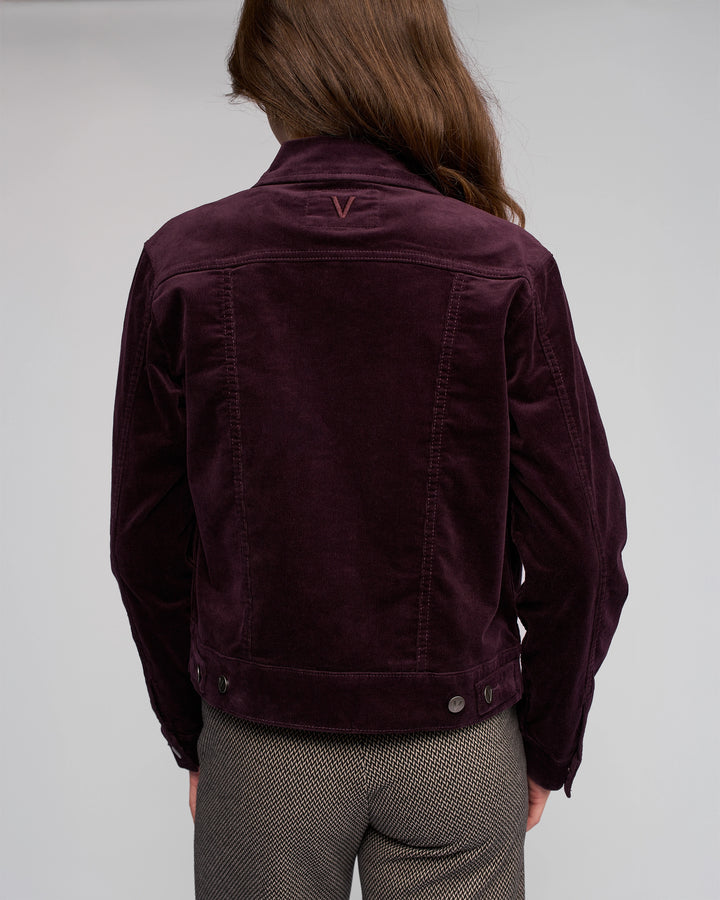 A person with long brown hair is seen from behind wearing the Vassalli Core Cord Jacket in mulberry, featuring a denim-jacket style with V stitching, paired with gray patterned pants against a plain light gray background.