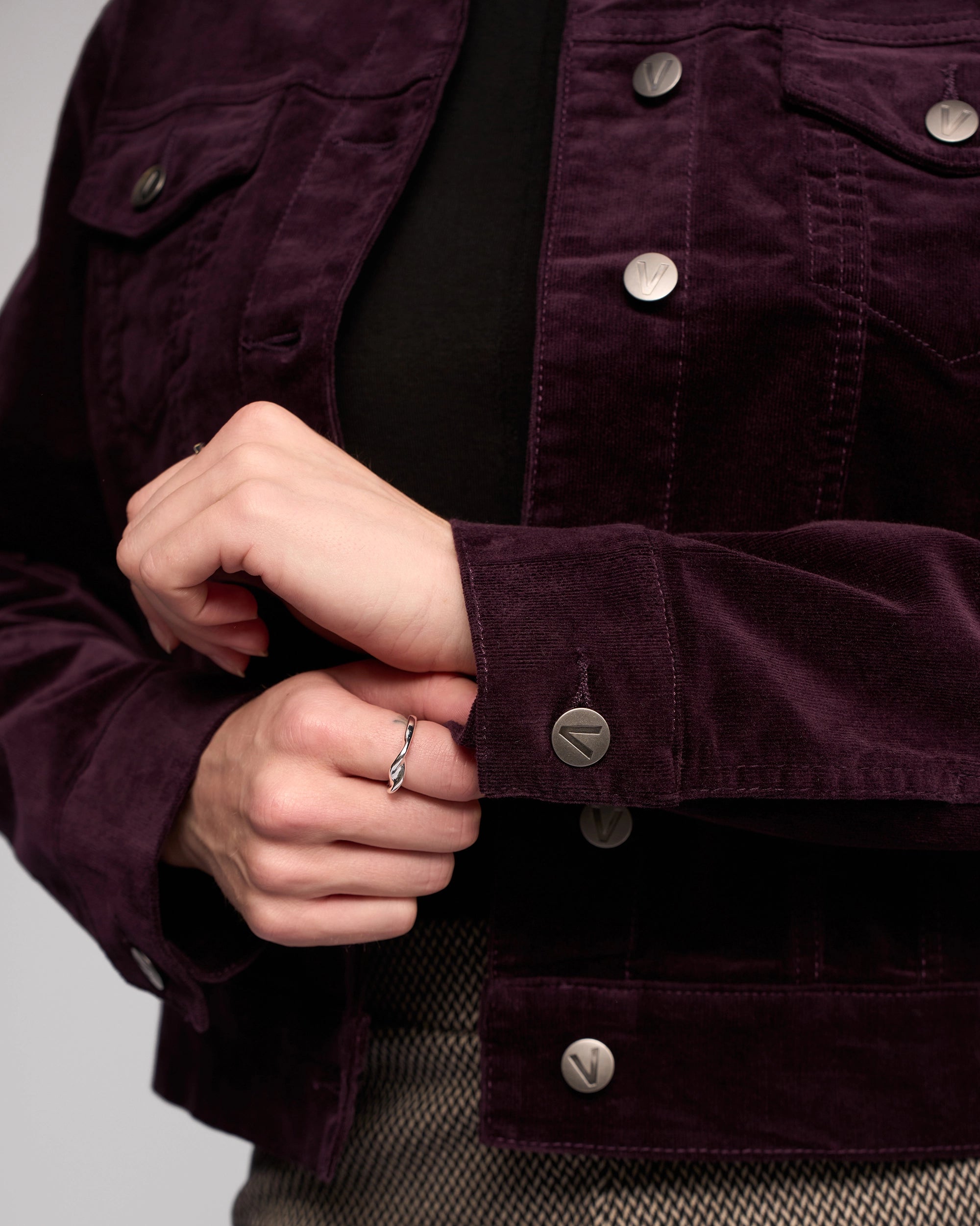 Close-up of a person buttoning the cuff of the Vassalli Core Cord Jacket in Mulberry, styled denim-jacket style. They're wearing a black top, light brown patterned skirt, and a simple ring on one hand.