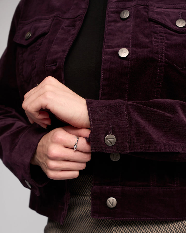 Close-up of a person buttoning the cuff of the Vassalli Core Cord Jacket in Mulberry, styled denim-jacket style. They're wearing a black top, light brown patterned skirt, and a simple ring on one hand.