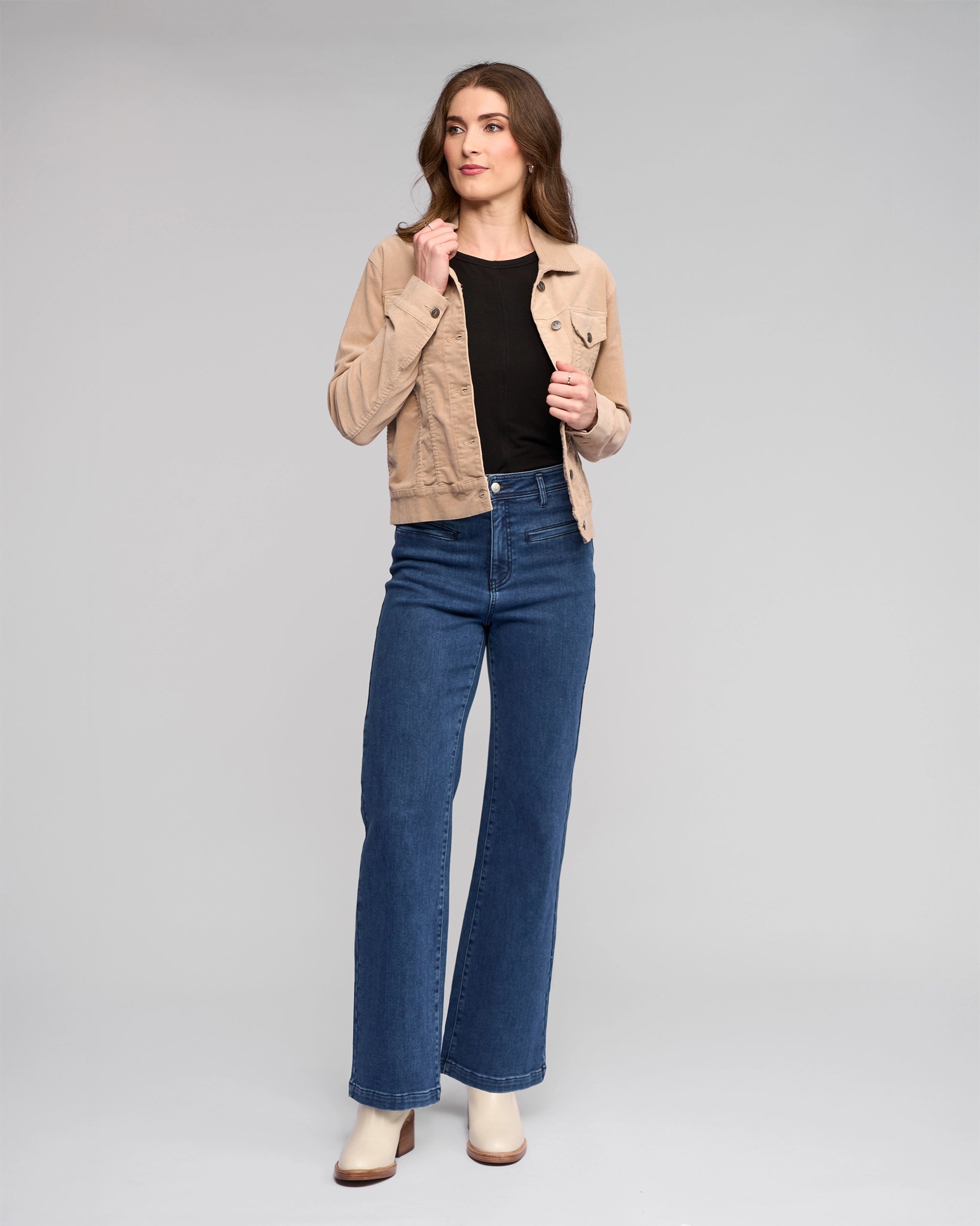 A woman with long brown hair poses confidently against a gray background, wearing the Vassalli Core Cord Jacket in Oat over a black top, blue high-waisted wide-leg jeans, and beige heeled boots.