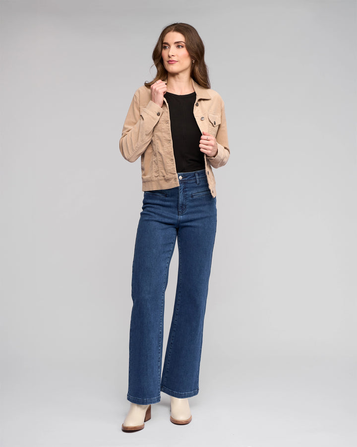A woman with long brown hair poses confidently against a gray background, wearing the Vassalli Core Cord Jacket in Oat over a black top, blue high-waisted wide-leg jeans, and beige heeled boots.