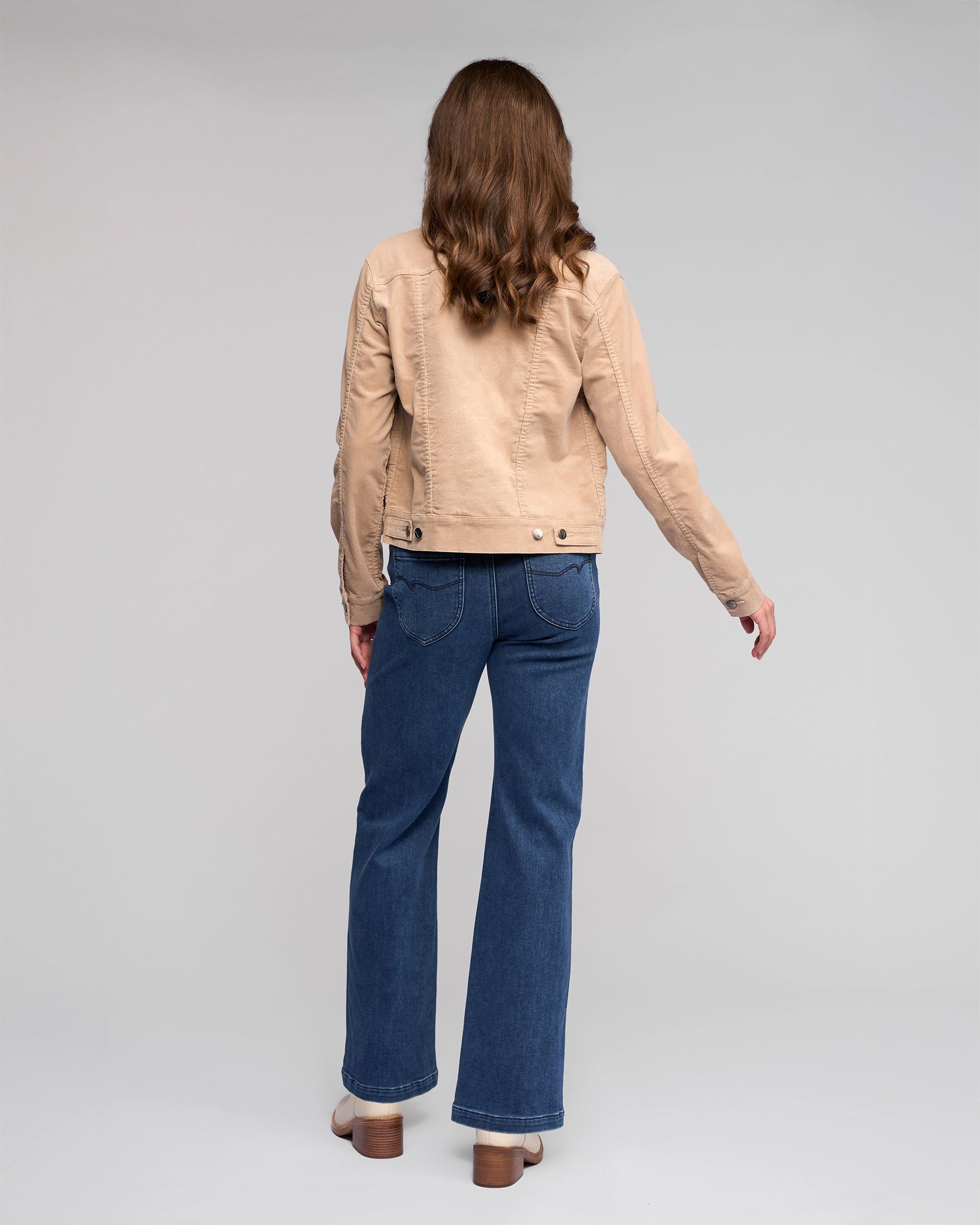A woman with wavy brown hair, wearing a Vassalli Core Cord Jacket in Oat, blue jeans, and heeled shoes, stands facing away from the camera against a plain gray background.