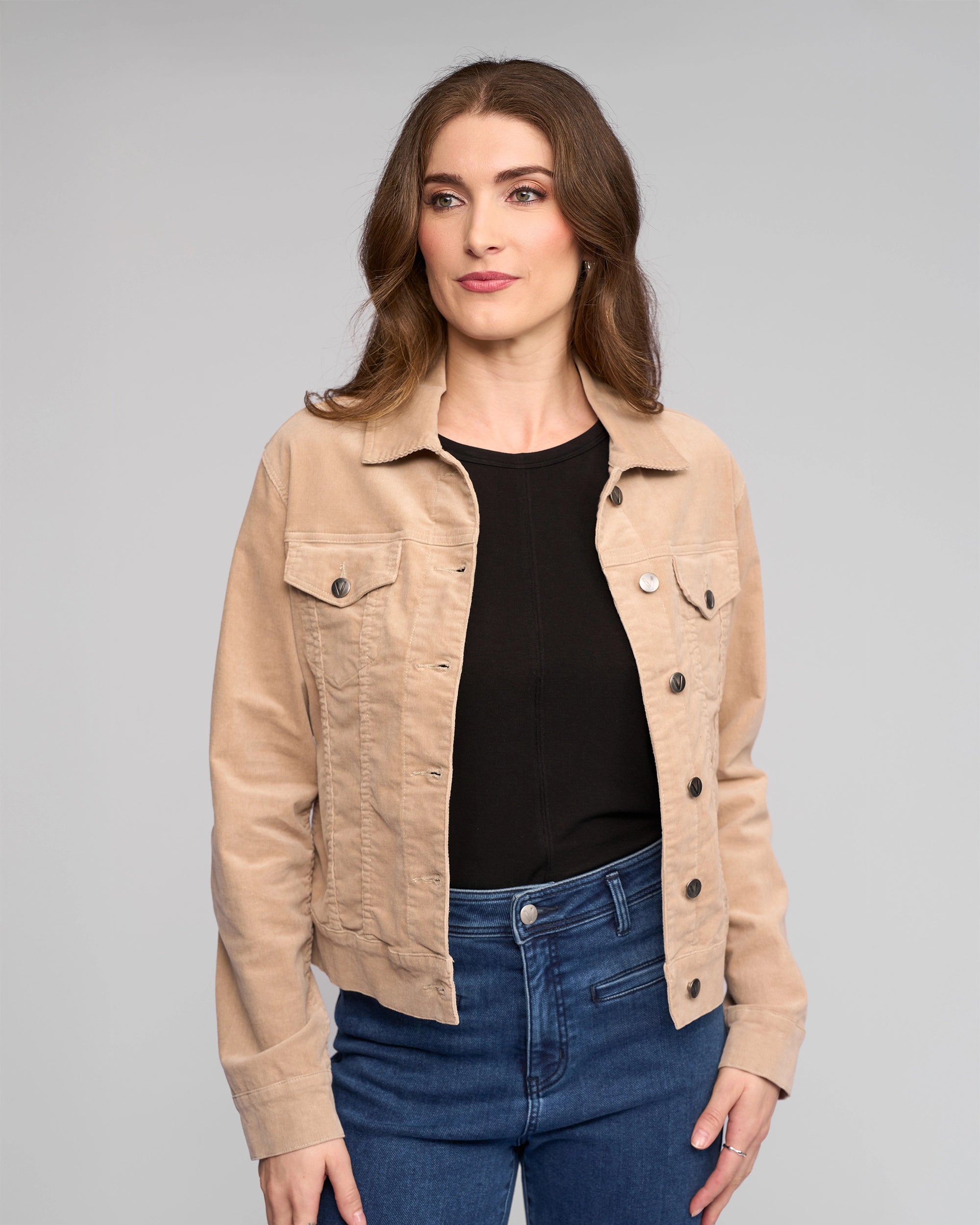 A woman with wavy brown hair wears the Vassalli Core Cord Jacket in Oat over a black top and blue jeans, standing against a plain light gray background and looking slightly to the side with a neutral expression.