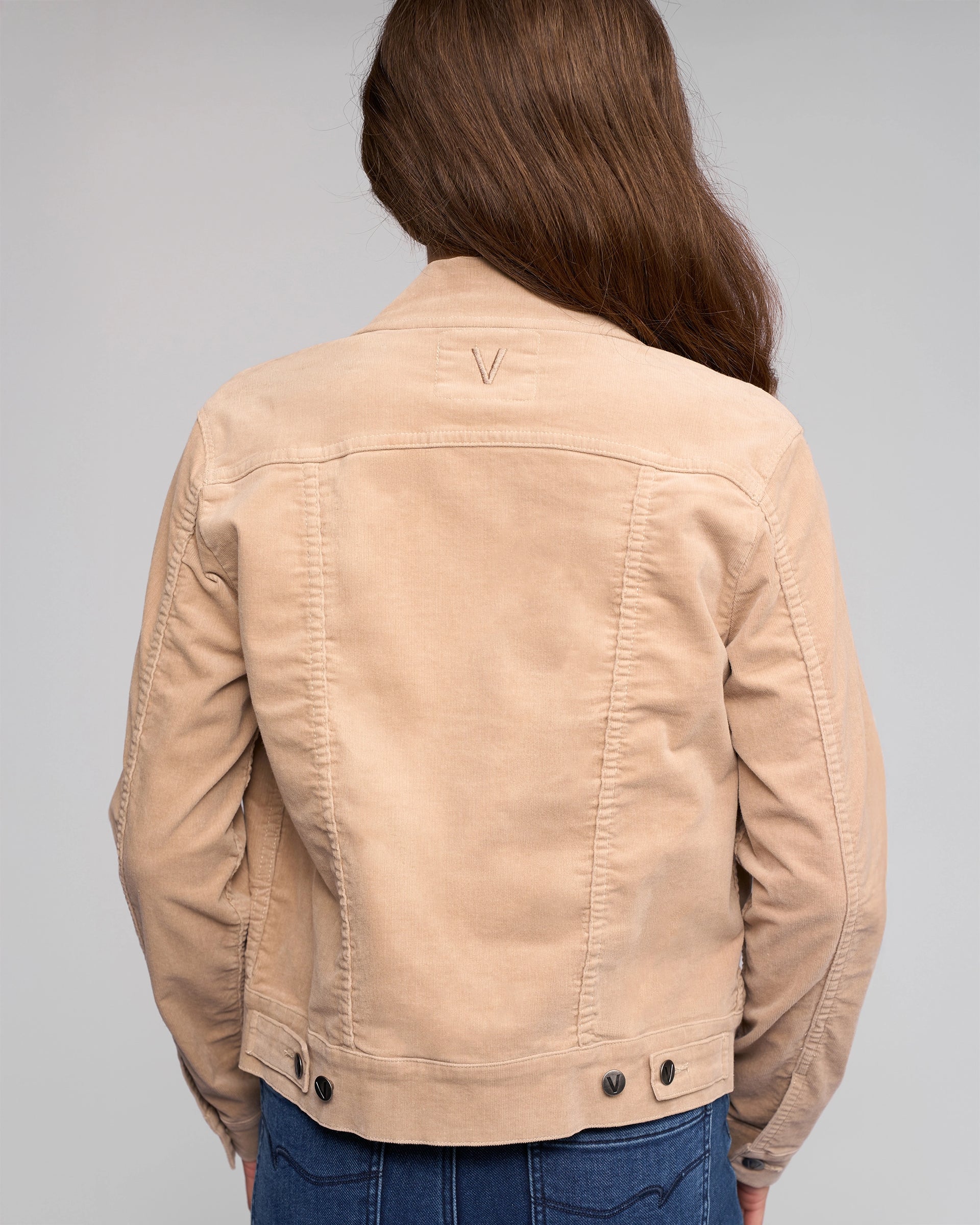 A person with long brown hair is seen from the back wearing a Vassalli Core Cord Jacket - Oat in a denim-jacket style and blue jeans, standing against a plain gray background.