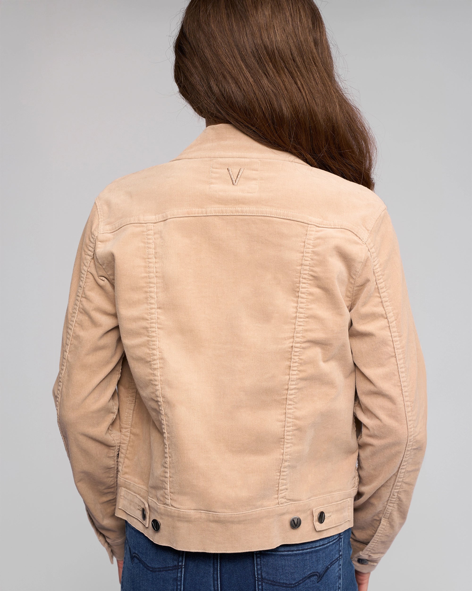 A person with long brown hair is seen from the back wearing a Vassalli Core Cord Jacket - Oat in a denim-jacket style and blue jeans, standing against a plain gray background.