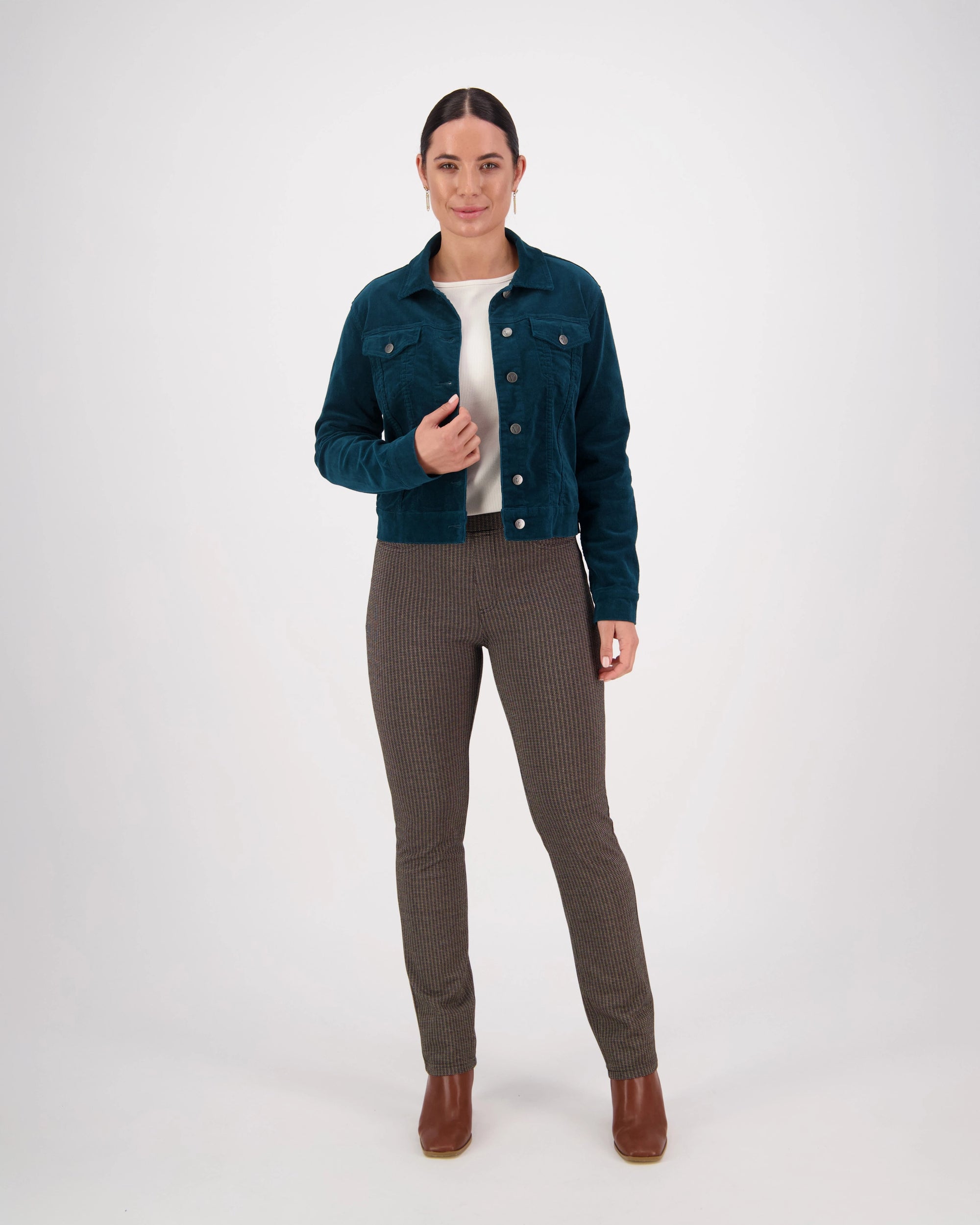 A woman with dark hair pulled back wears the Vassalli Core Cord Jacket in Peacock, styled denim-jacket fashion, over a white top, brown plaid pants, and brown ankle boots. She stands against a plain white background with a neutral expression.