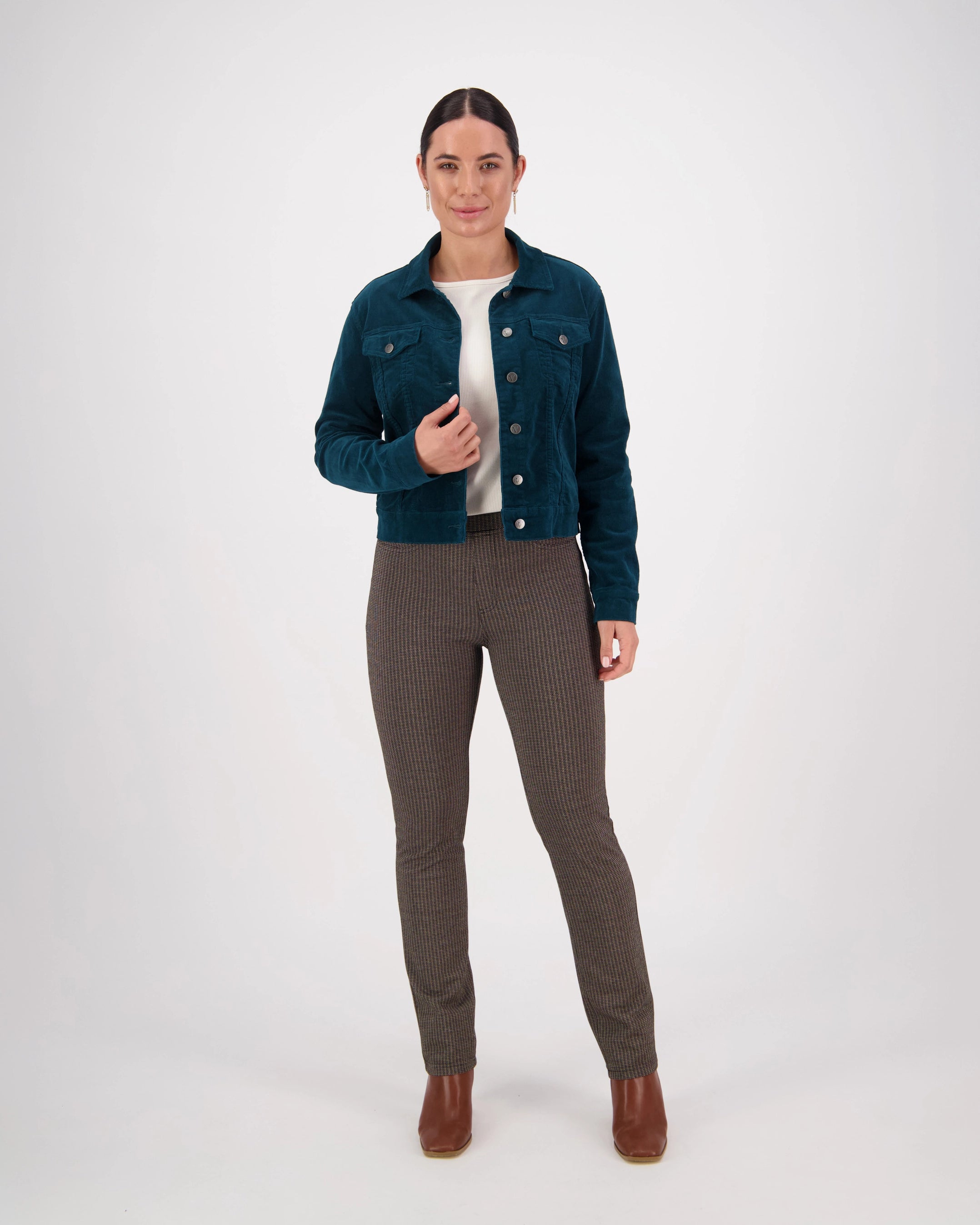 A woman with dark hair pulled back wears the Vassalli Core Cord Jacket in Peacock, styled denim-jacket fashion, over a white top, brown plaid pants, and brown ankle boots. She stands against a plain white background with a neutral expression.