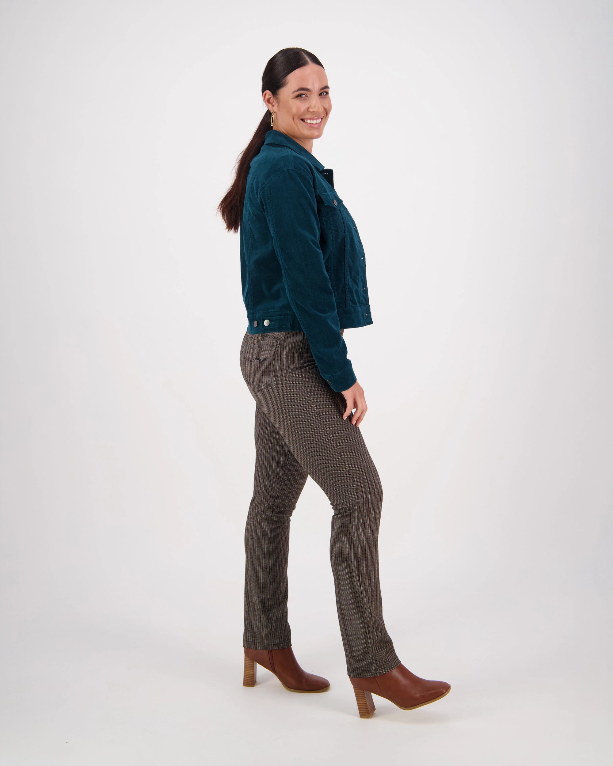 A woman with long dark hair in a ponytail smiles while wearing the Vassalli Core Cord Jacket in Peacock, brown checkered pants, and brown heeled boots, posing sideways against a plain white background.