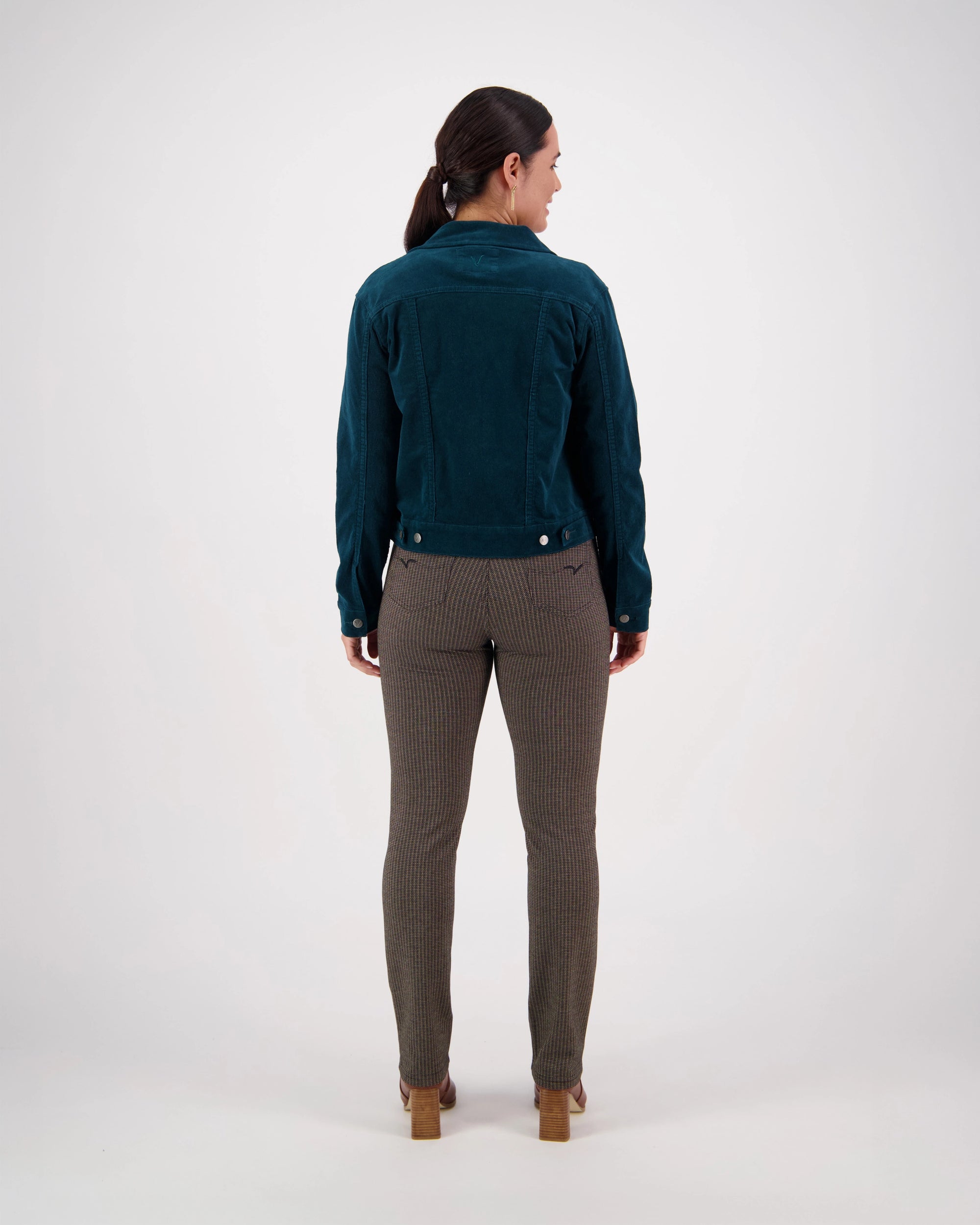 A woman faces away from the camera, wearing the Vassalli Core Cord Jacket in Peacock, brown checkered pants, and tan heeled shoes against a plain white background.