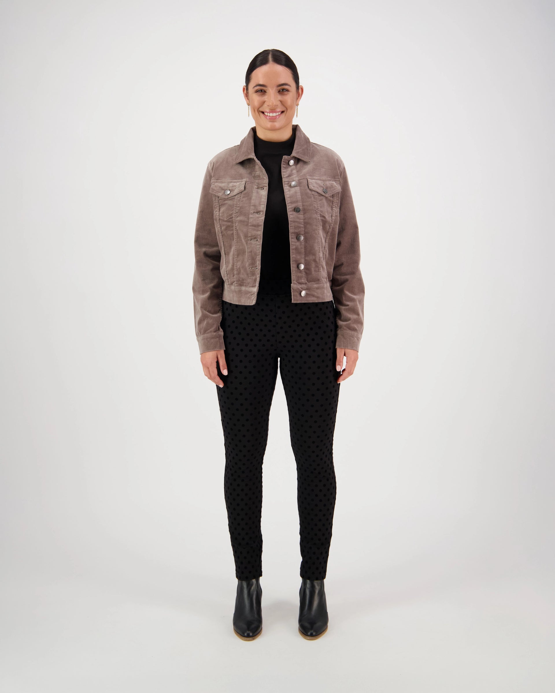 A woman faces forward, smiling, in a Vassalli Core Cord Jacket - Taupe styled like a denim jacket, paired with a black turtleneck, dotted black pants, and black ankle boots. The background is plain and light-colored.
