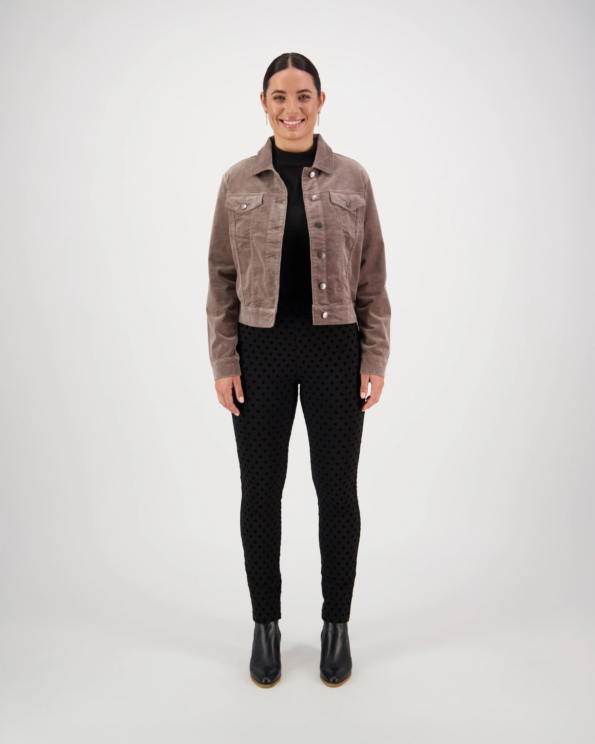 A woman faces forward, smiling, in a Vassalli Core Cord Jacket - Taupe styled like a denim jacket, paired with a black turtleneck, dotted black pants, and black ankle boots. The background is plain and light-colored.