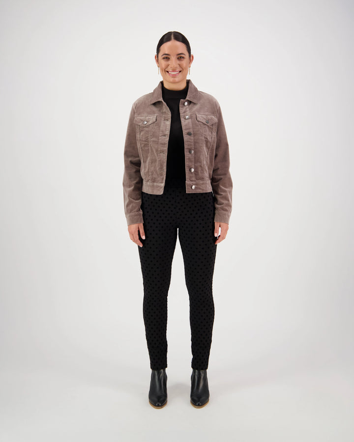 A woman faces forward, smiling, in a Vassalli Core Cord Jacket - Taupe styled like a denim jacket, paired with a black turtleneck, dotted black pants, and black ankle boots. The background is plain and light-colored.