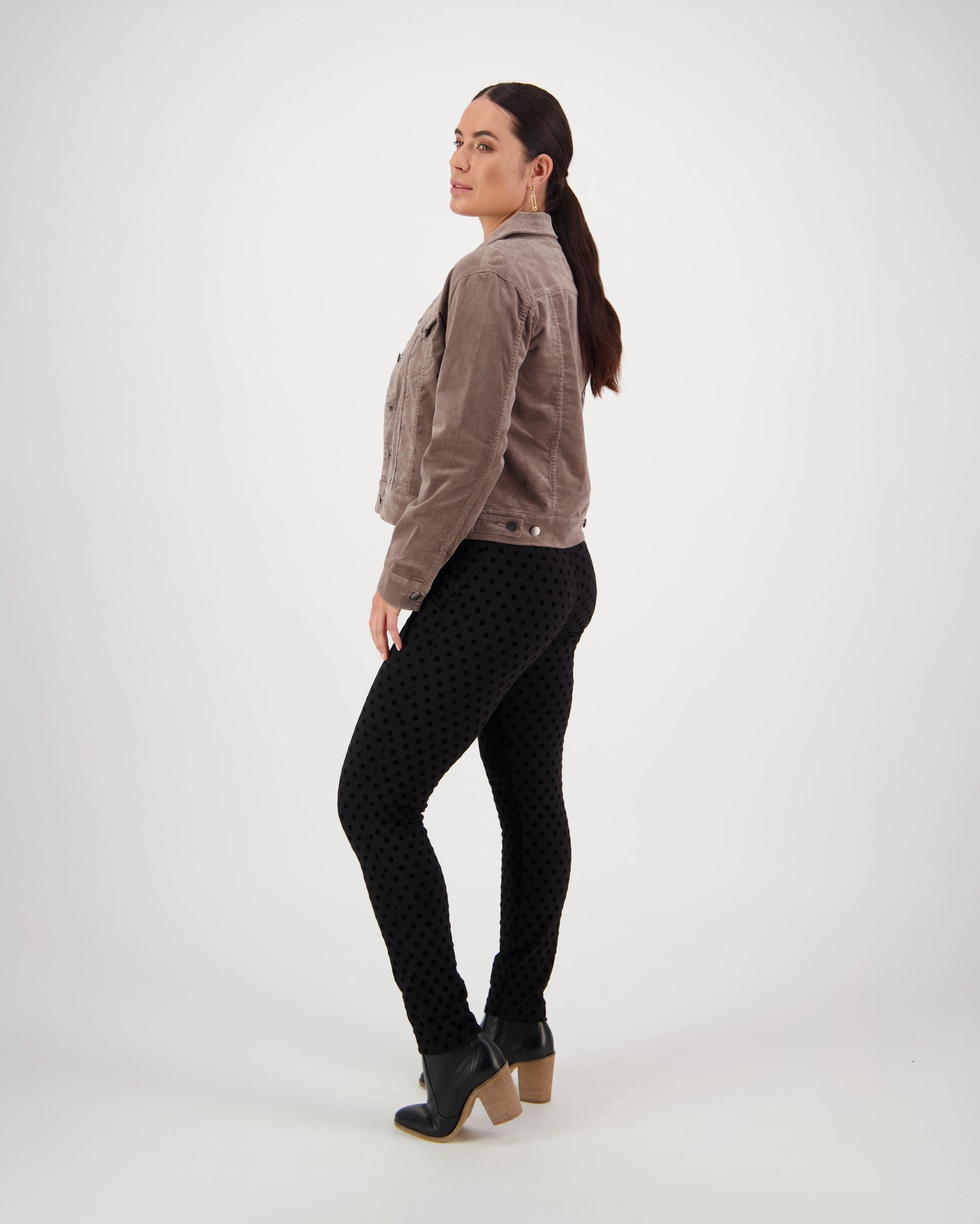A woman with dark hair in a ponytail stands sideways, looking at the camera. She wears the Vassalli Core Cord Jacket in taupe, black polka-dot pants, and black ankle boots with tan heels against a plain white background.