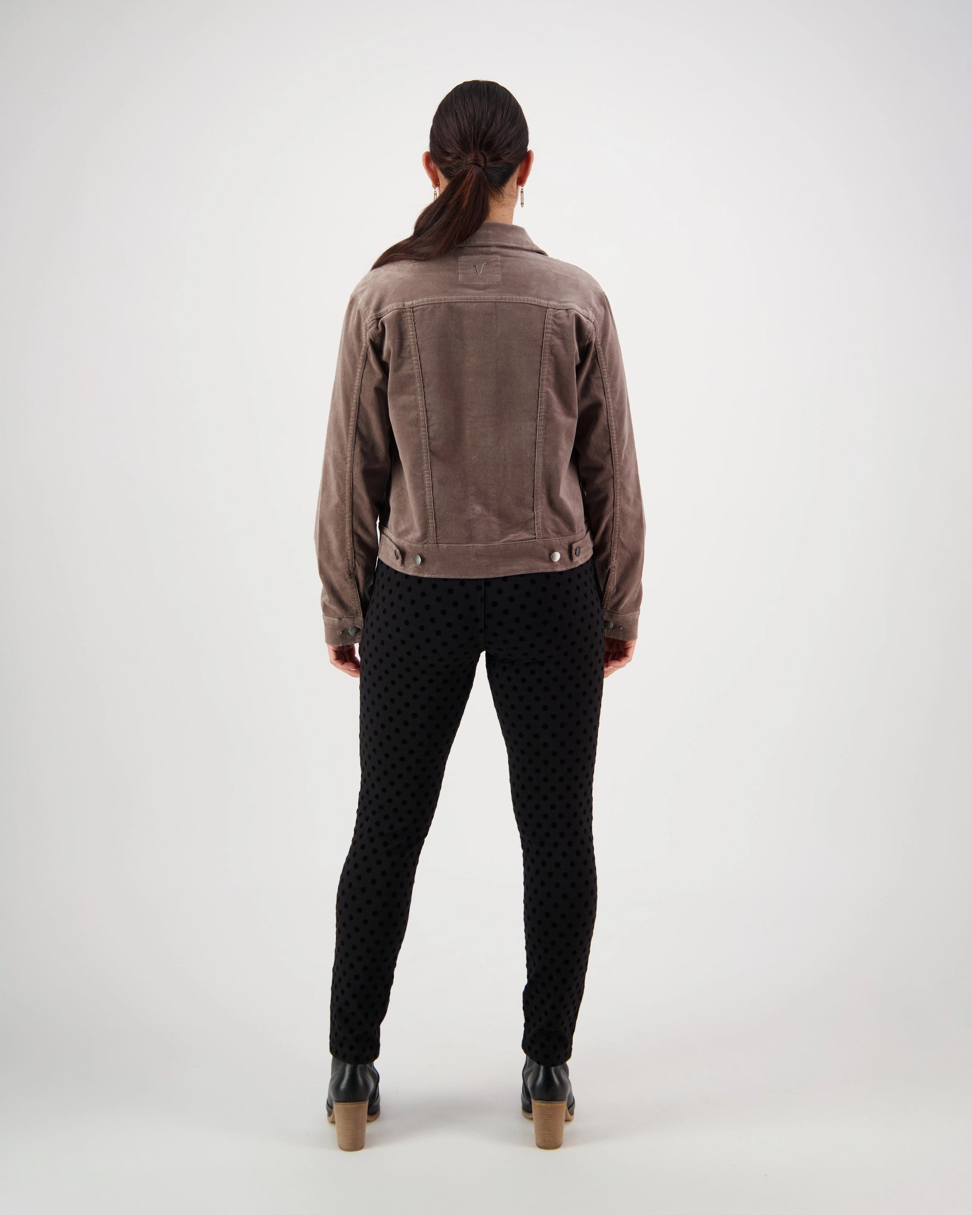 A person with long dark hair tied back, wearing the Vassalli Core Cord Jacket in taupe, black textured pants, and heeled shoes stands facing away on a plain white background.
