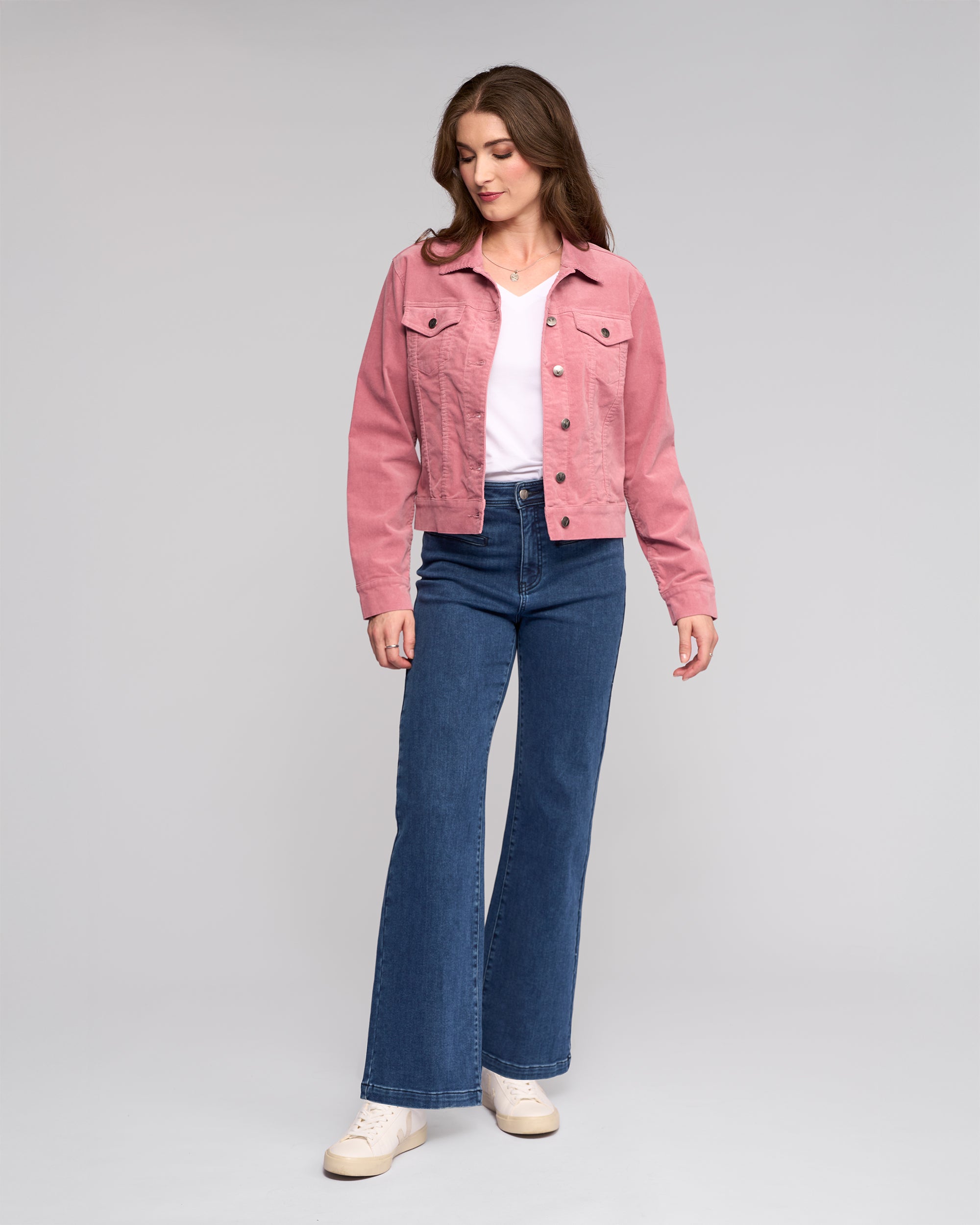 A woman with long brown hair stands against a plain background, wearing the Vassalli Core Cord Jacket in dusky pink over a white top, blue high-waisted jeans, and white sneakers. She looks slightly down to her left in a relaxed pose.