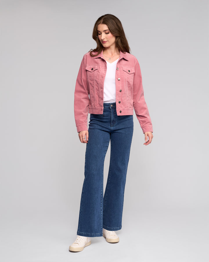 A woman with long brown hair stands against a plain background, wearing the Vassalli Core Cord Jacket in dusky pink over a white top, blue high-waisted jeans, and white sneakers. She looks slightly down to her left in a relaxed pose.