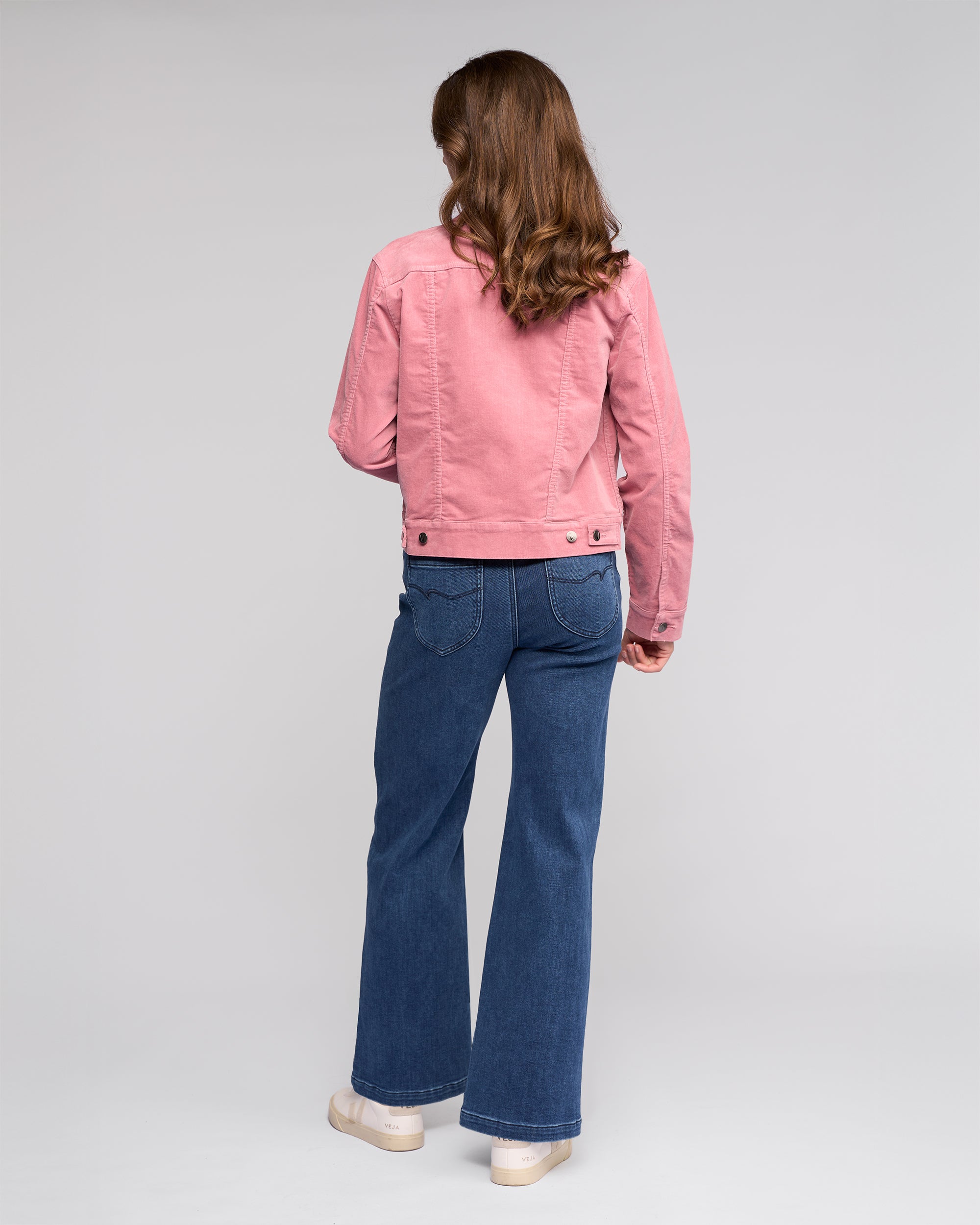 A person with long, wavy brown hair faces away, wearing the Vassalli Core Cord Jacket in Dusky Pink with blue jeans and white shoes against a plain light background.