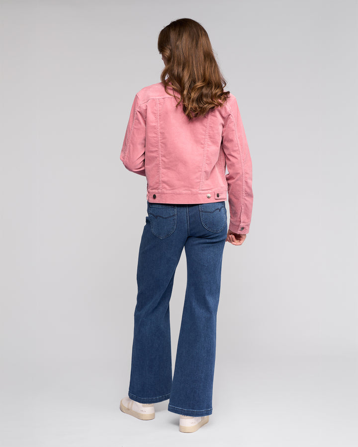 A person with long, wavy brown hair faces away, wearing the Vassalli Core Cord Jacket in Dusky Pink with blue jeans and white shoes against a plain light background.