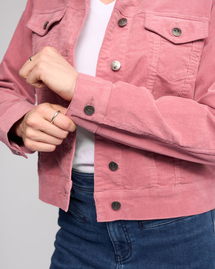 A person wearing the Vassalli Core Cord Jacket in Dusky Pink over a white shirt and blue jeans adjusts the jacket's sleeve. Only their upper body and hands are visible.