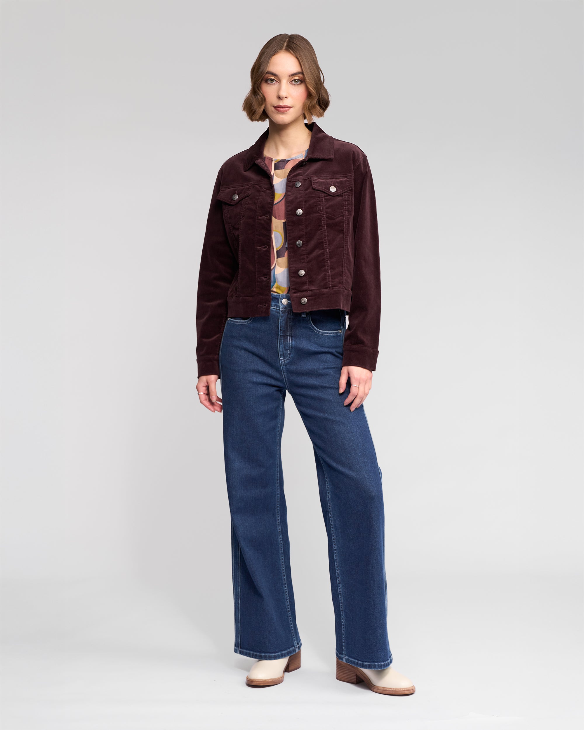 A woman stands facing forward, wearing the Vassalli Core Cord Jacket in Merlot, a printed top, wide-leg blue jeans, and beige boots against a plain light gray background.