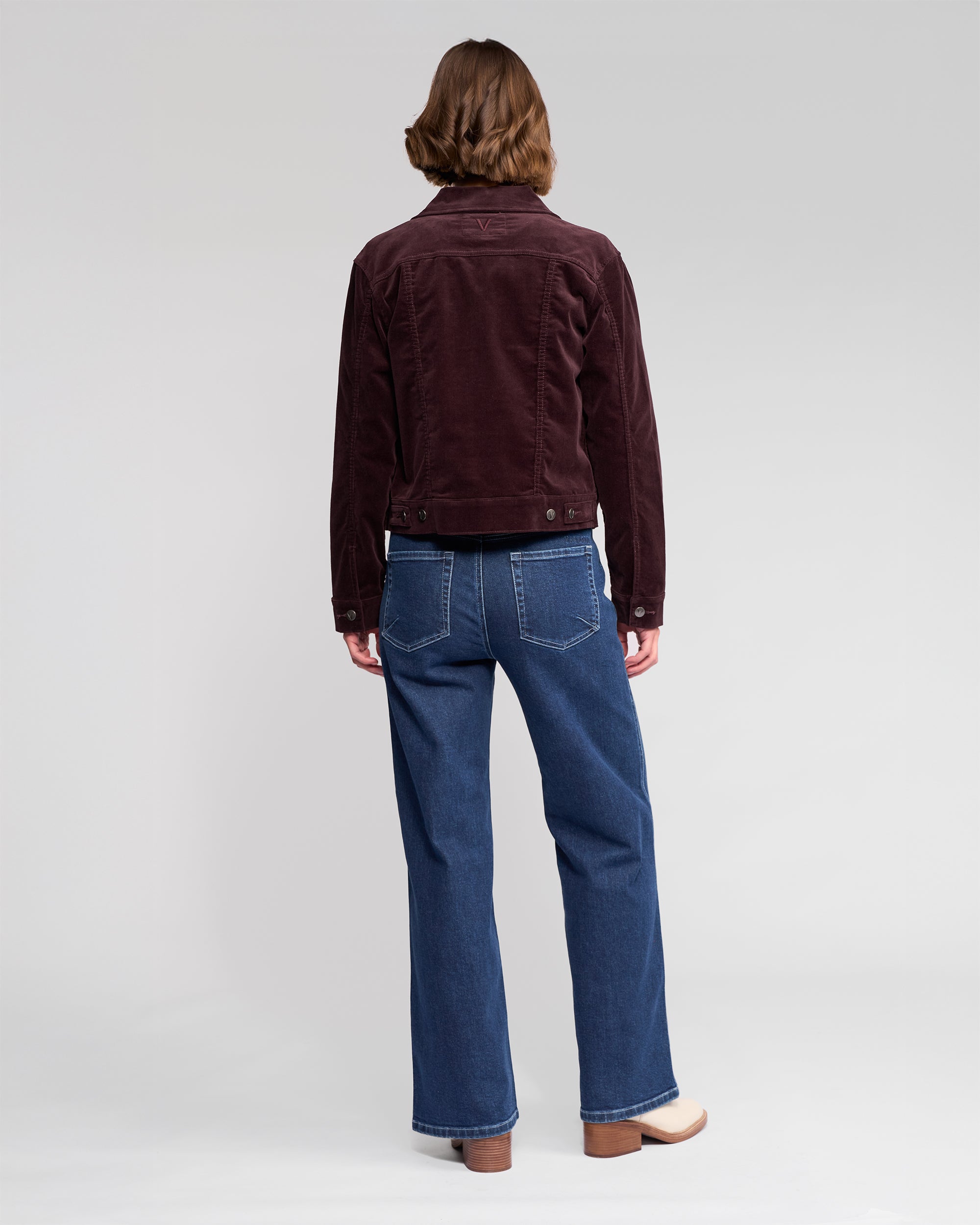 A person stands with their back to the camera, wearing the Vassalli Core Cord Jacket in Merlot, blue jeans featuring visible stitching, and light-colored shoes against a plain light background.