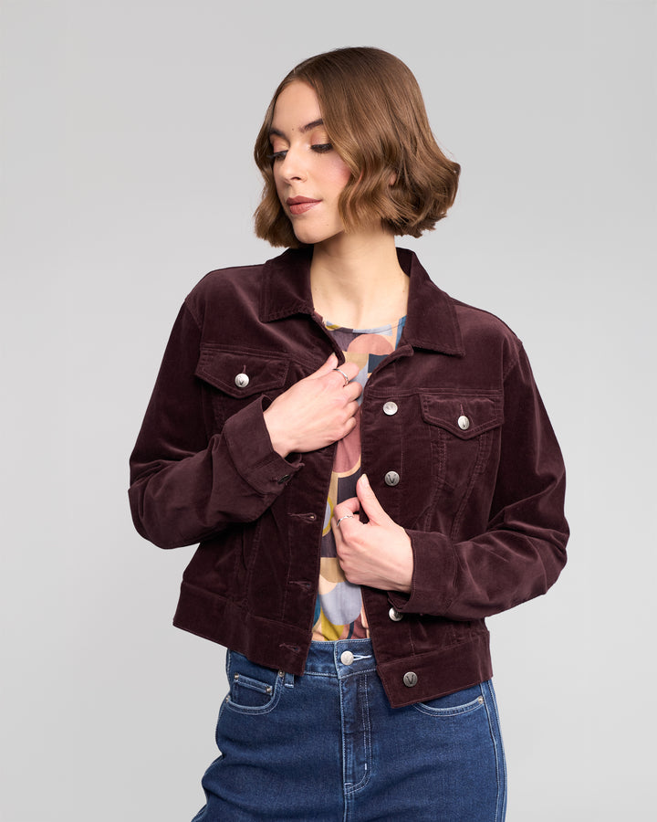A woman with short brown hair wears the Vassalli Core Cord Jacket in Merlot over a colorful top and blue jeans, standing against a plain light gray background, looking down and slightly to the side.