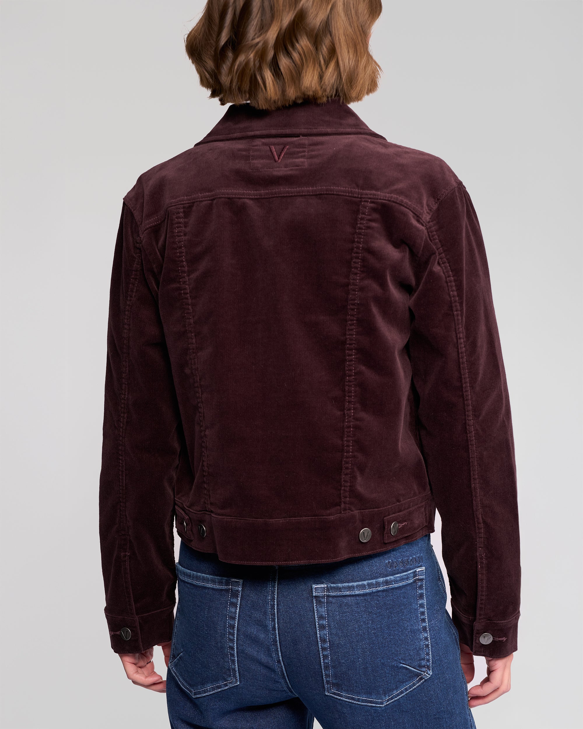 A person with shoulder-length brown hair is shown from the back wearing the Vassalli Core Cord Jacket in Merlot and blue jeans against a plain, light gray background.
