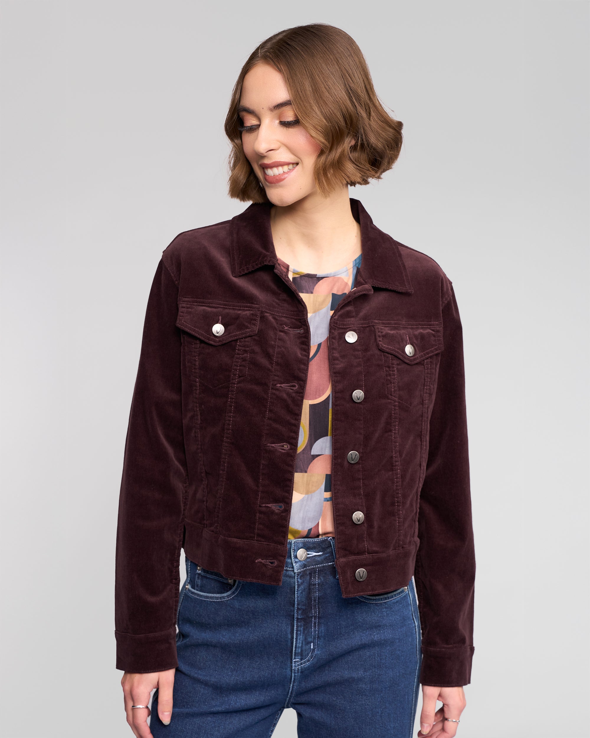 A woman with short brown hair smiles while looking down, wearing the Vassalli Core Cord Jacket in Merlot over a colorful patterned blouse and blue jeans, standing against a light gray background.