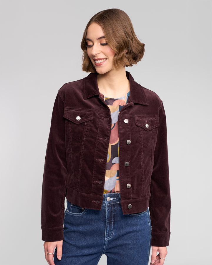 A woman with short brown hair smiles while looking down, wearing the Vassalli Core Cord Jacket in Merlot over a colorful patterned blouse and blue jeans, standing against a light gray background.