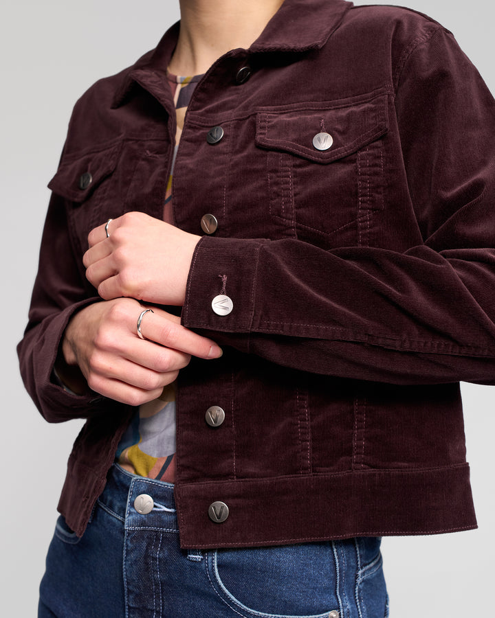 A person wears the Vassalli Core Cord Jacket in Merlot with metal buttons and blue jeans, shown from shoulders to hips. Their hands are lightly clasped at the front, displaying a silver ring.
