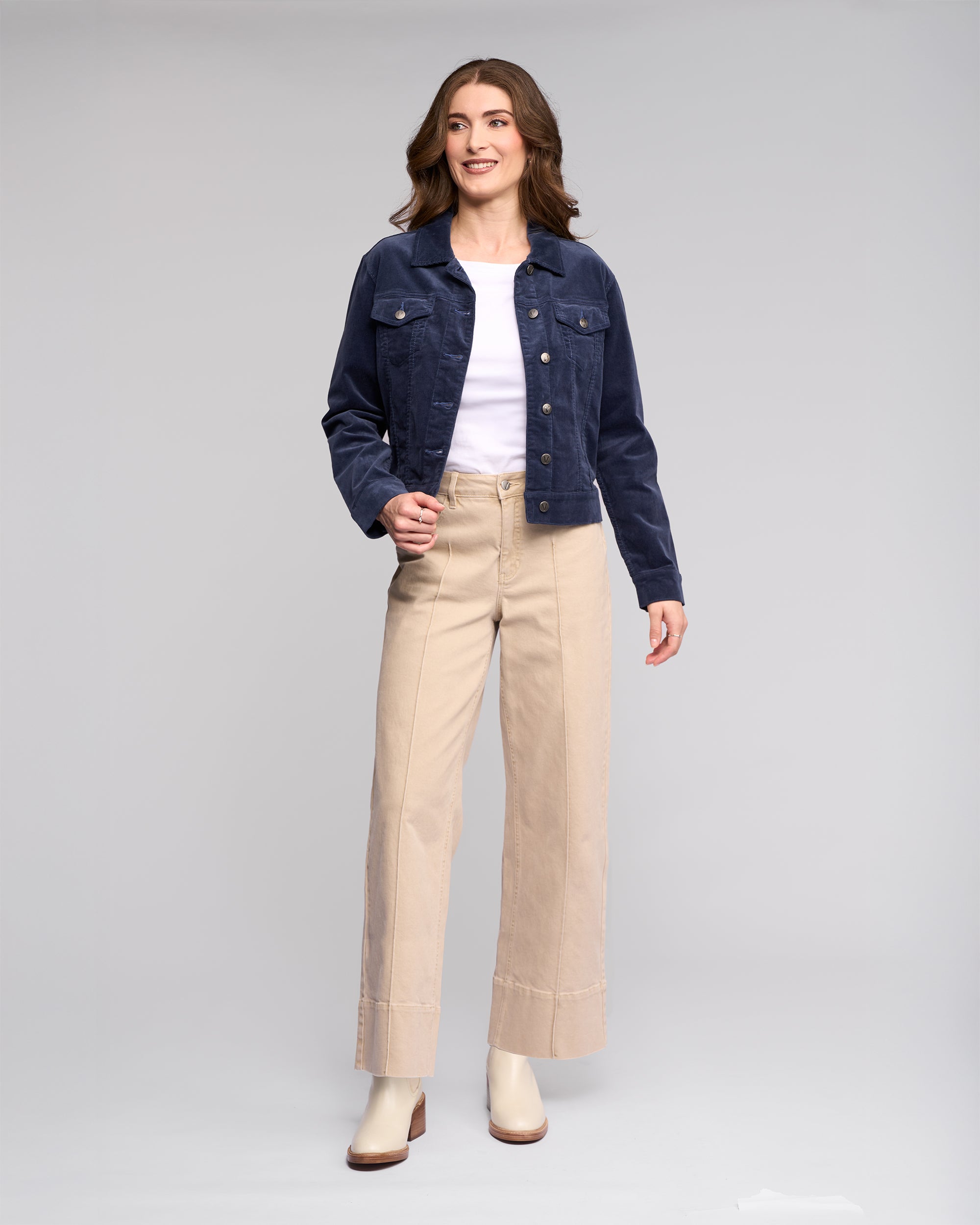 A woman smiles, wearing the Vassalli Core Cord Jacket in Slate over a white shirt, beige wide-leg pants, and light ankle boots, standing against a plain light gray background.