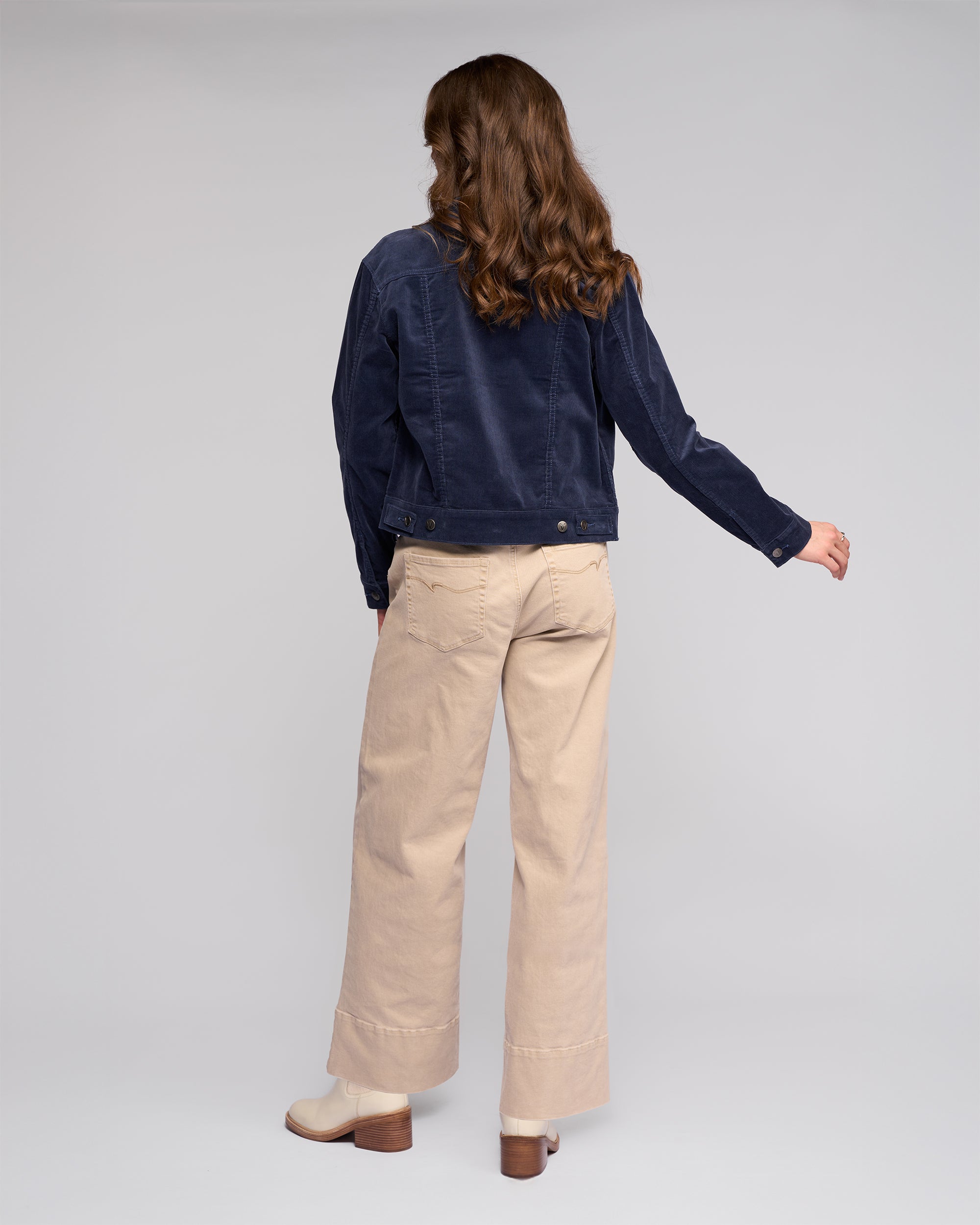 A person with wavy brown hair stands facing away, wearing the Vassalli Core Cord Jacket in Slate, beige wide-leg pants, and light heeled boots against a plain light gray background.