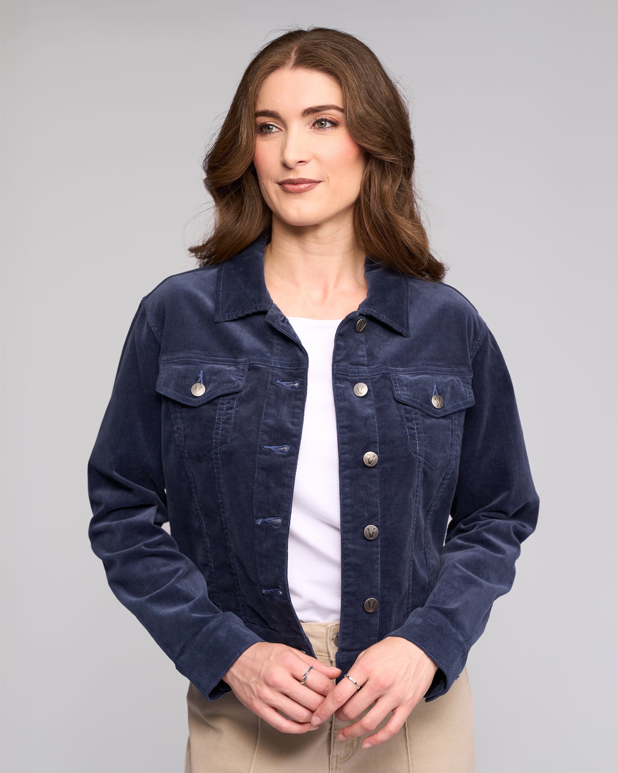 A woman with wavy brown hair models the Vassalli Core Cord Jacket in Slate over a white shirt and beige pants, standing against a plain light gray background.