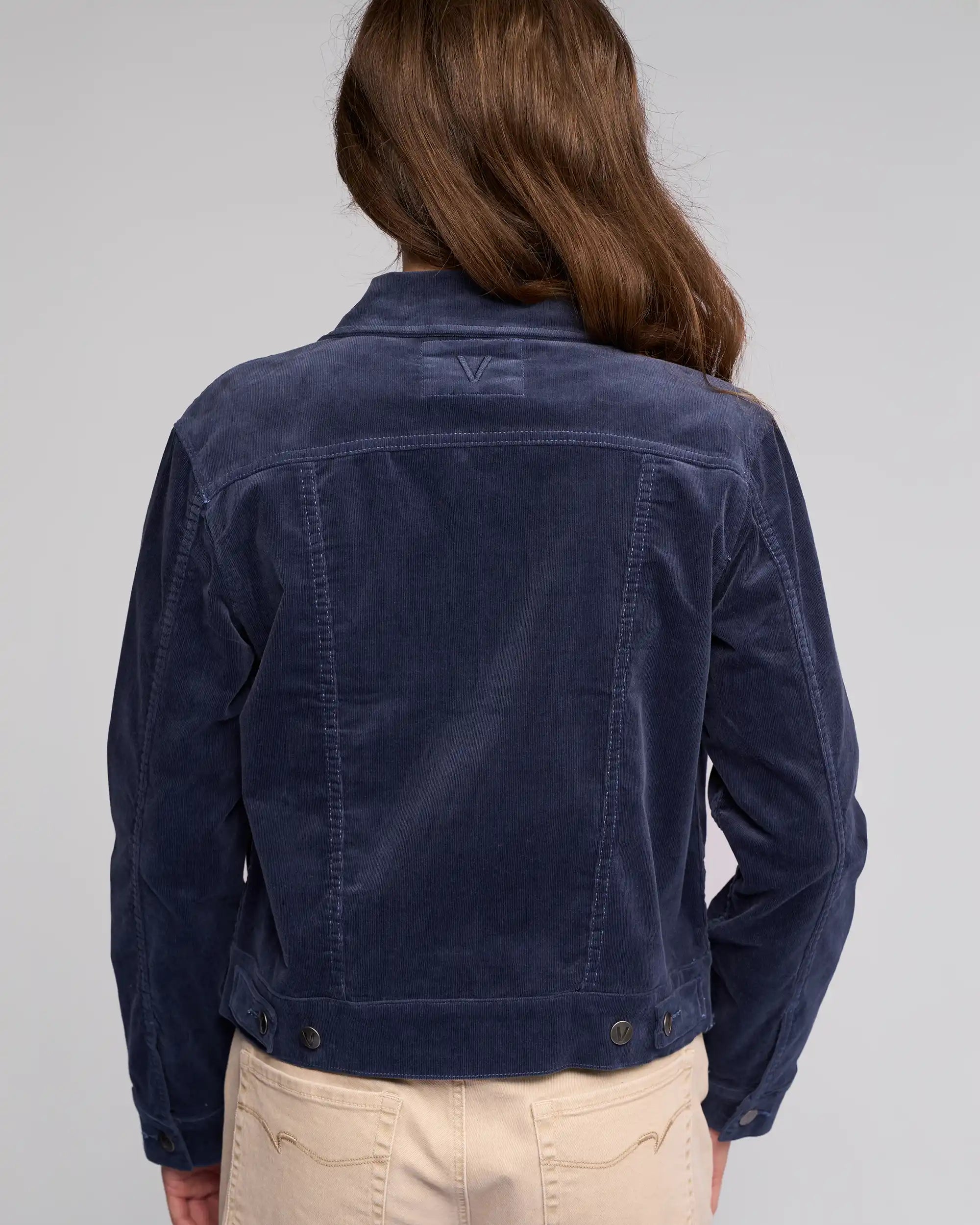 A person with long brown hair is shown from the back, wearing the Vassalli Core Cord Jacket in Slate and beige pants, against a plain light gray background.