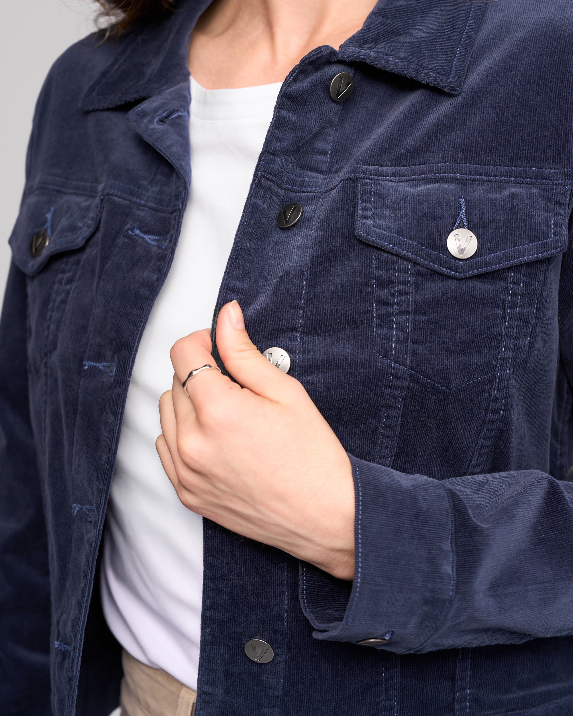 A person is shown from the shoulders down, adjusting a silver button on their Vassalli Core Cord Jacket in Slate, worn over a white shirt.