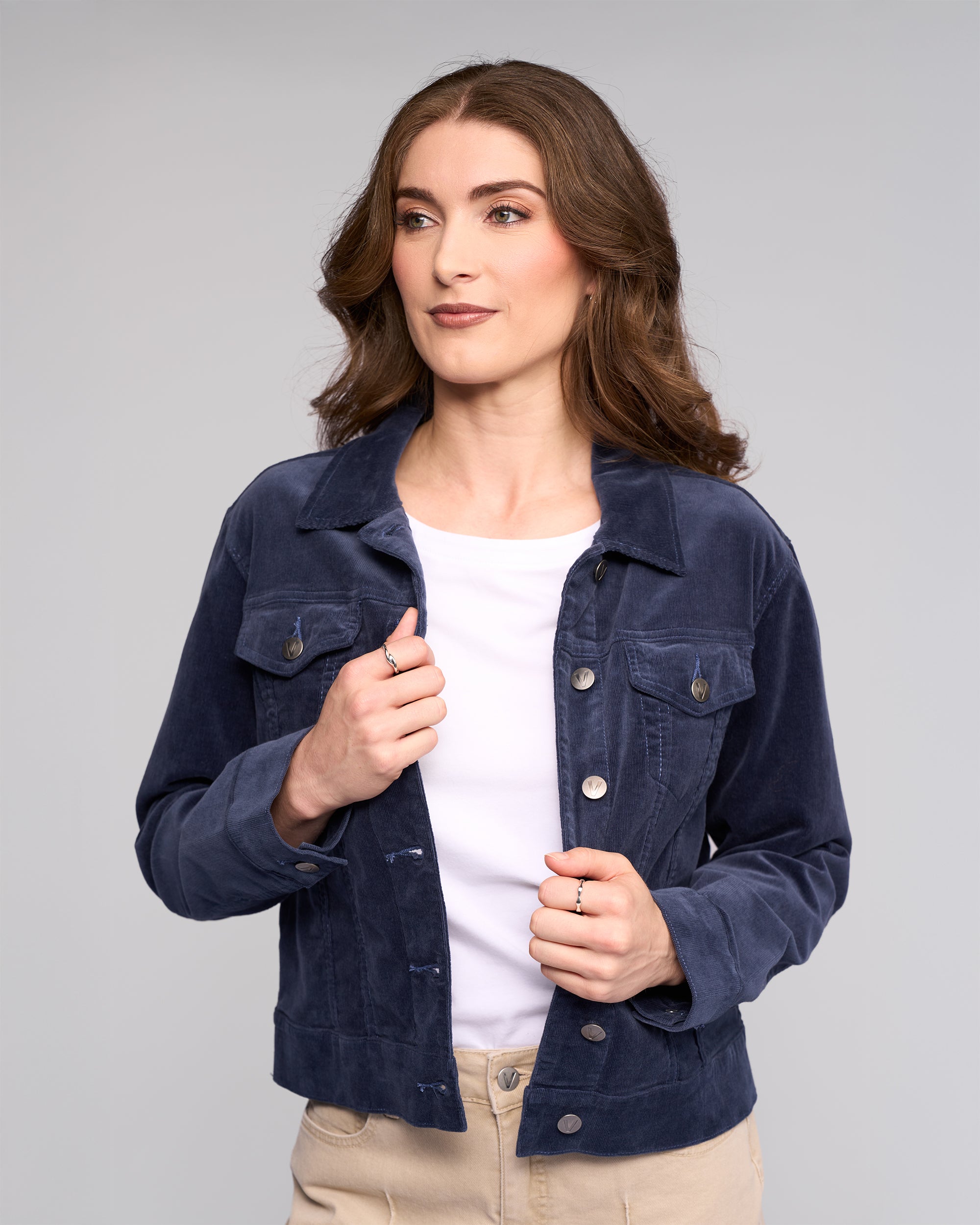 A woman with wavy brown hair wears the Vassalli Core Cord Jacket in Slate over a white shirt and beige pants, standing before a light gray background. She holds the jacket's edges and gazes slightly to the side.
