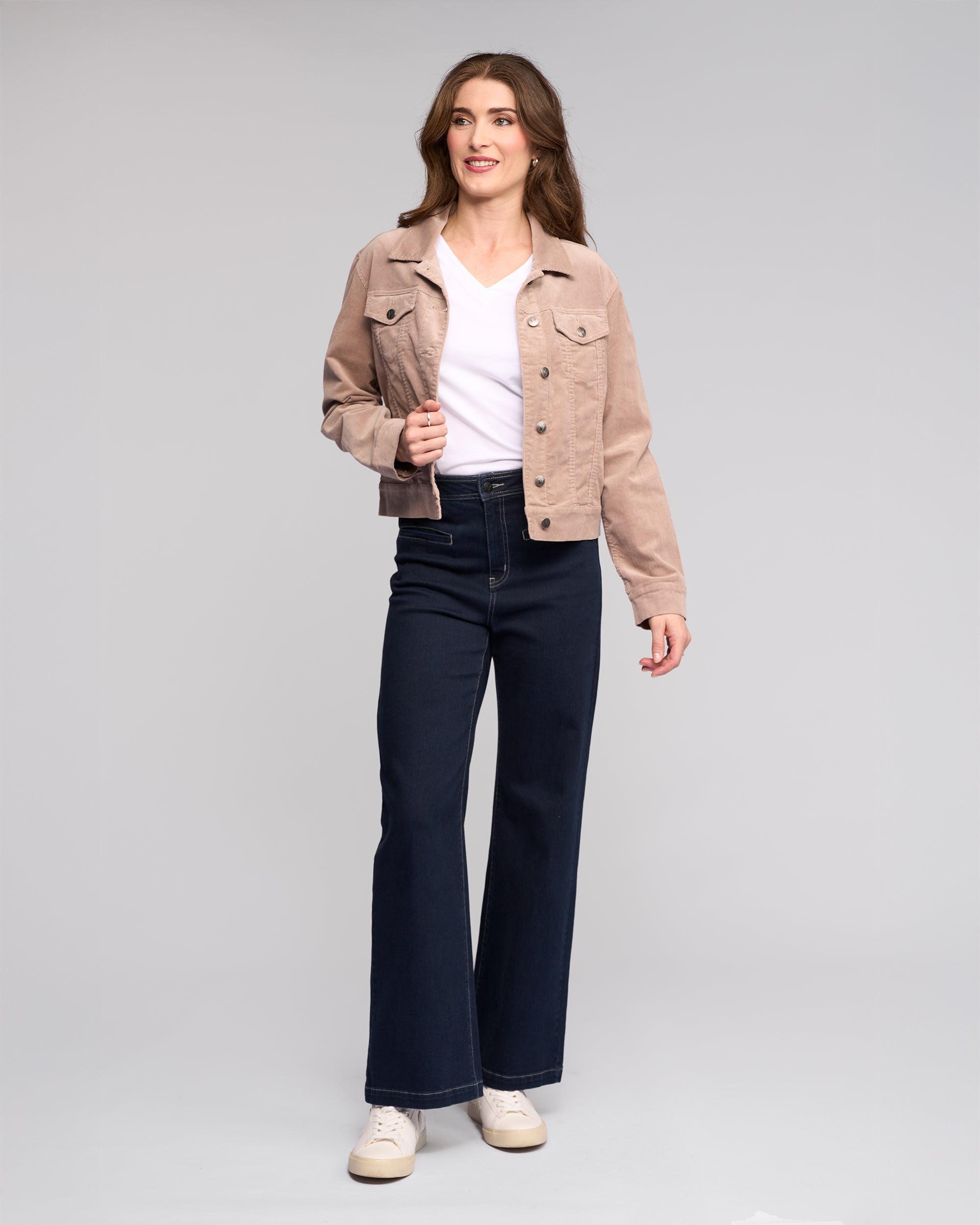 A woman with long brown hair stands against a plain background, smiling softly in the Vassalli Core Cord Jacket - Twig over a white top, paired with dark high-waisted wide-leg jeans and beige sneakers.
