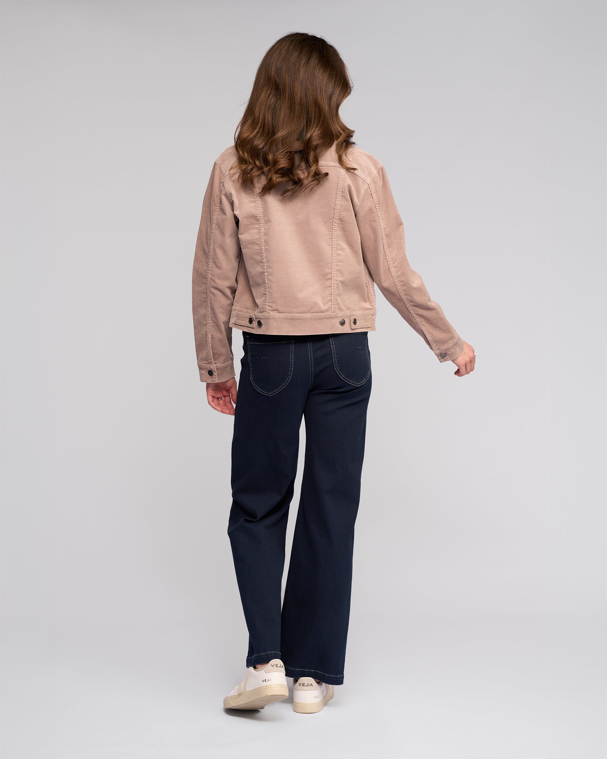A woman with wavy brown hair stands facing away, wearing the Vassalli Core Cord Jacket in Twig, paired with dark blue jeans and white shoes, set against a plain light gray background.