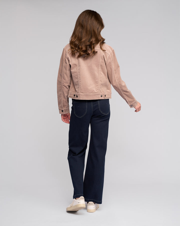 A woman with wavy brown hair stands facing away, wearing the Vassalli Core Cord Jacket in Twig, paired with dark blue jeans and white shoes, set against a plain light gray background.