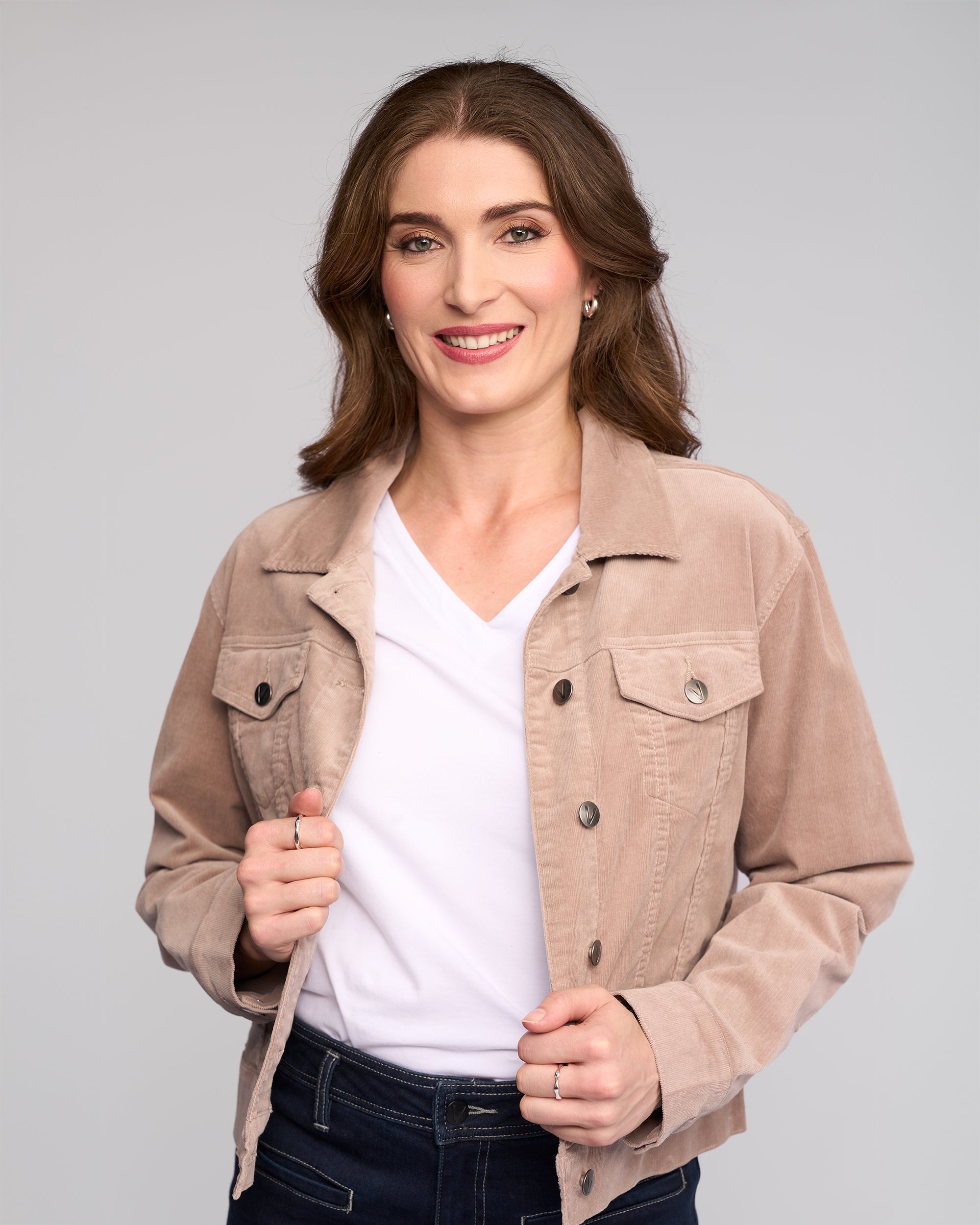 A woman with long brown hair smiles at the camera, wearing the Vassalli Core Cord Jacket in Twig over a white V-neck shirt and dark jeans, standing against a plain light gray background.