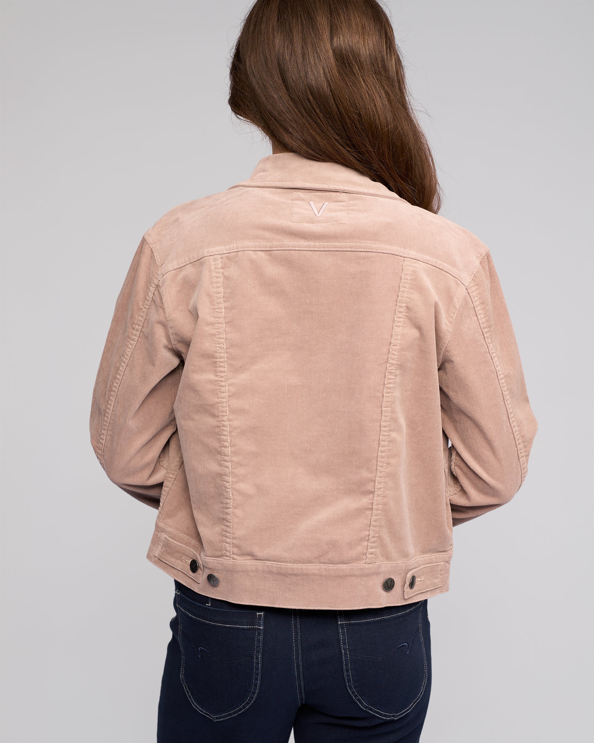 A woman with long brown hair, seen from behind, wears the Vassalli Core Cord Jacket in Twig and dark blue jeans against a plain gray background.