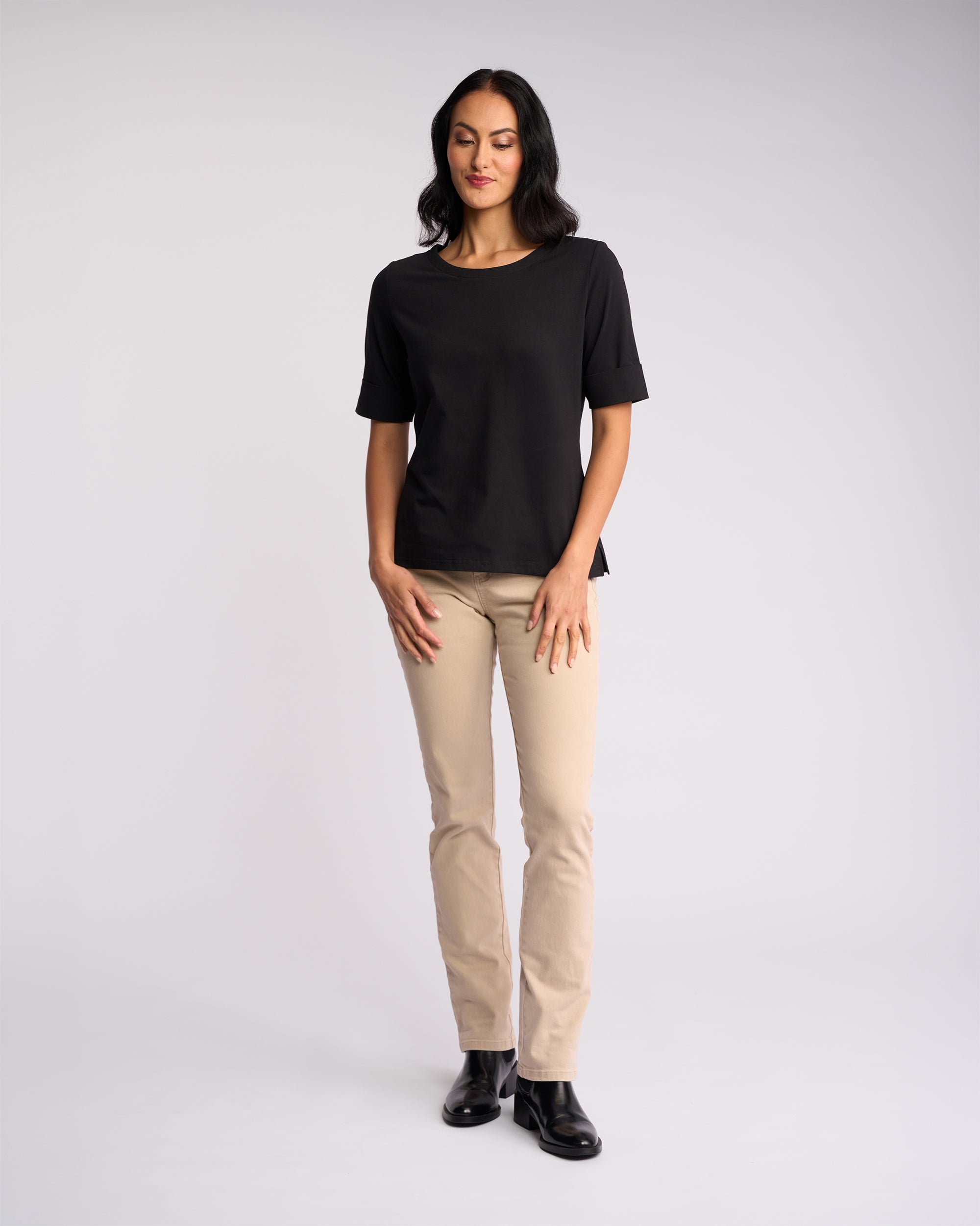A woman stands against a light background, smiling softly and wearing the Vassalli Arden Top in black with short sleeves, paired with beige pants and black ankle boots. Her hands rest relaxed by her sides.