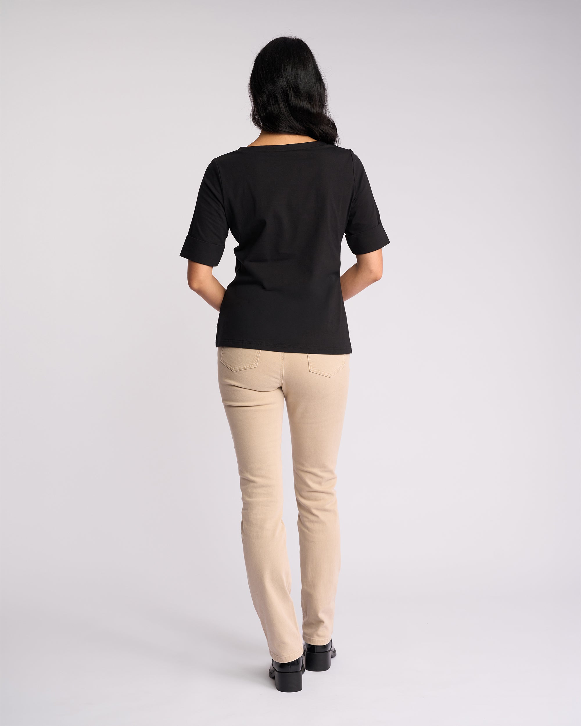 A person with long dark hair stands facing away, wearing the Vassalli Arden Top in black, beige pants, and black shoes against a plain light background.