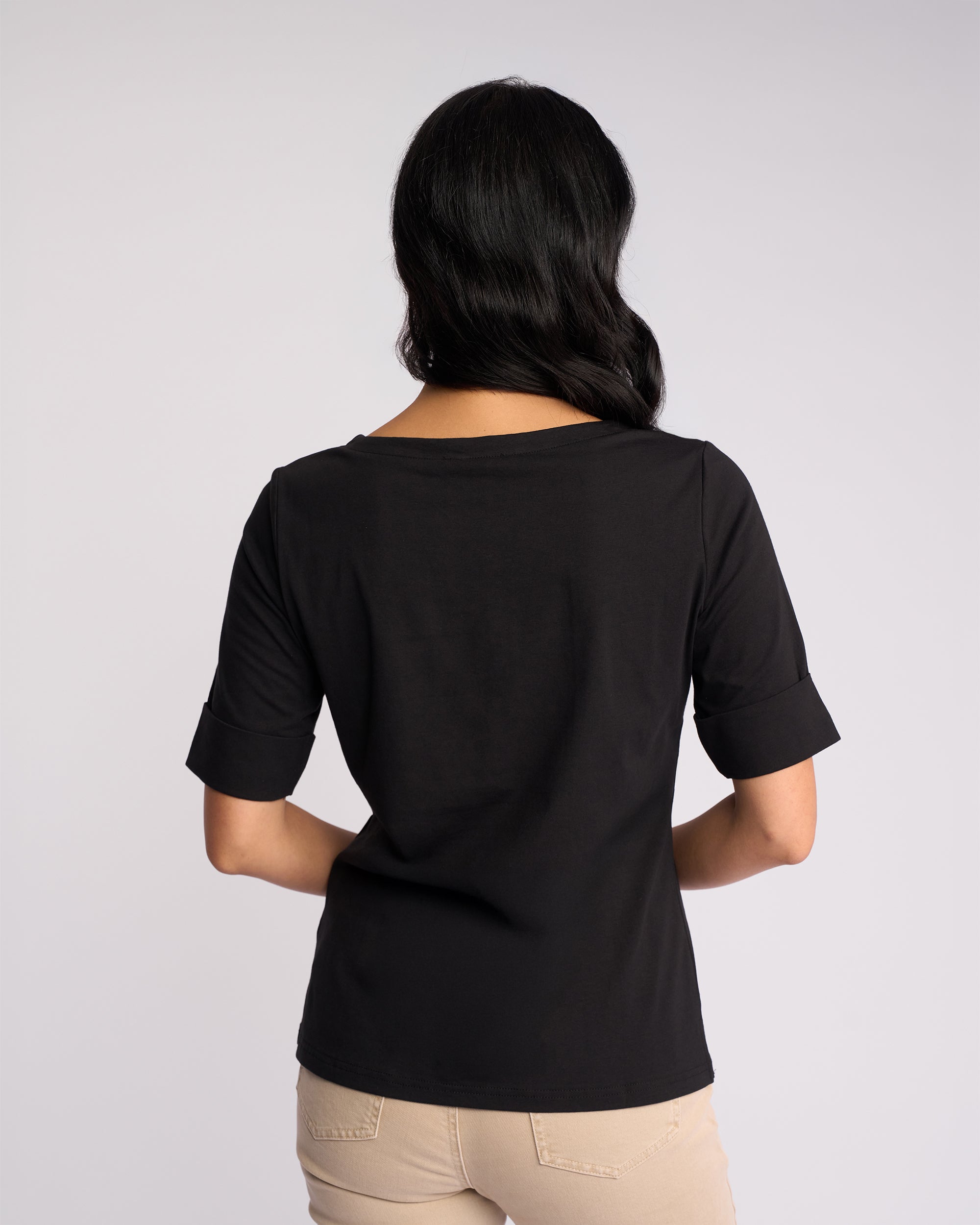 A woman with long dark hair stands facing away, wearing the Vassalli Arden Top in black and beige pants against a light gray background.