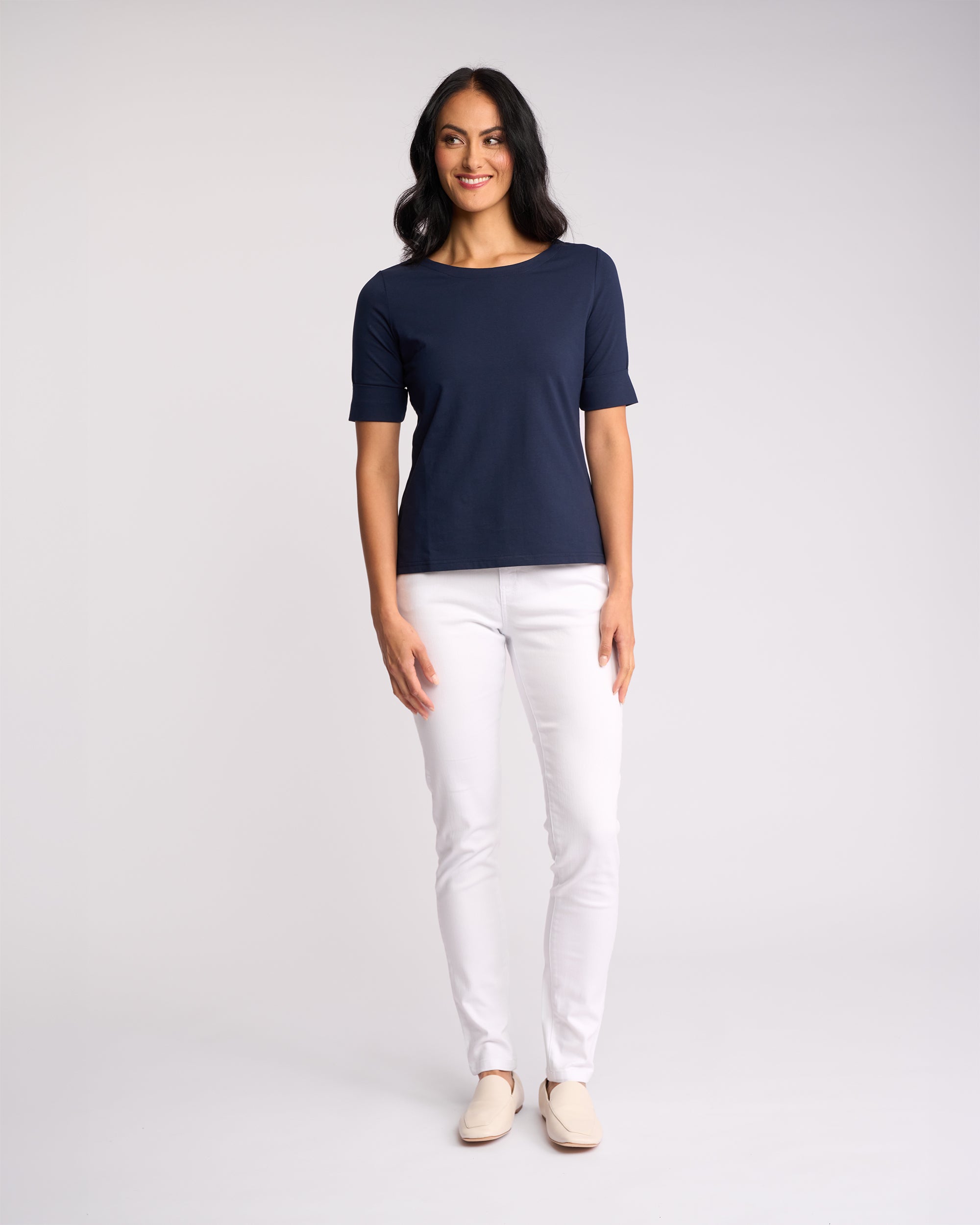 A woman smiles against a plain light background, wearing the Vassalli Arden Top in navy with a boat neck and stretch fabric, styled with white pants and cream shoes.