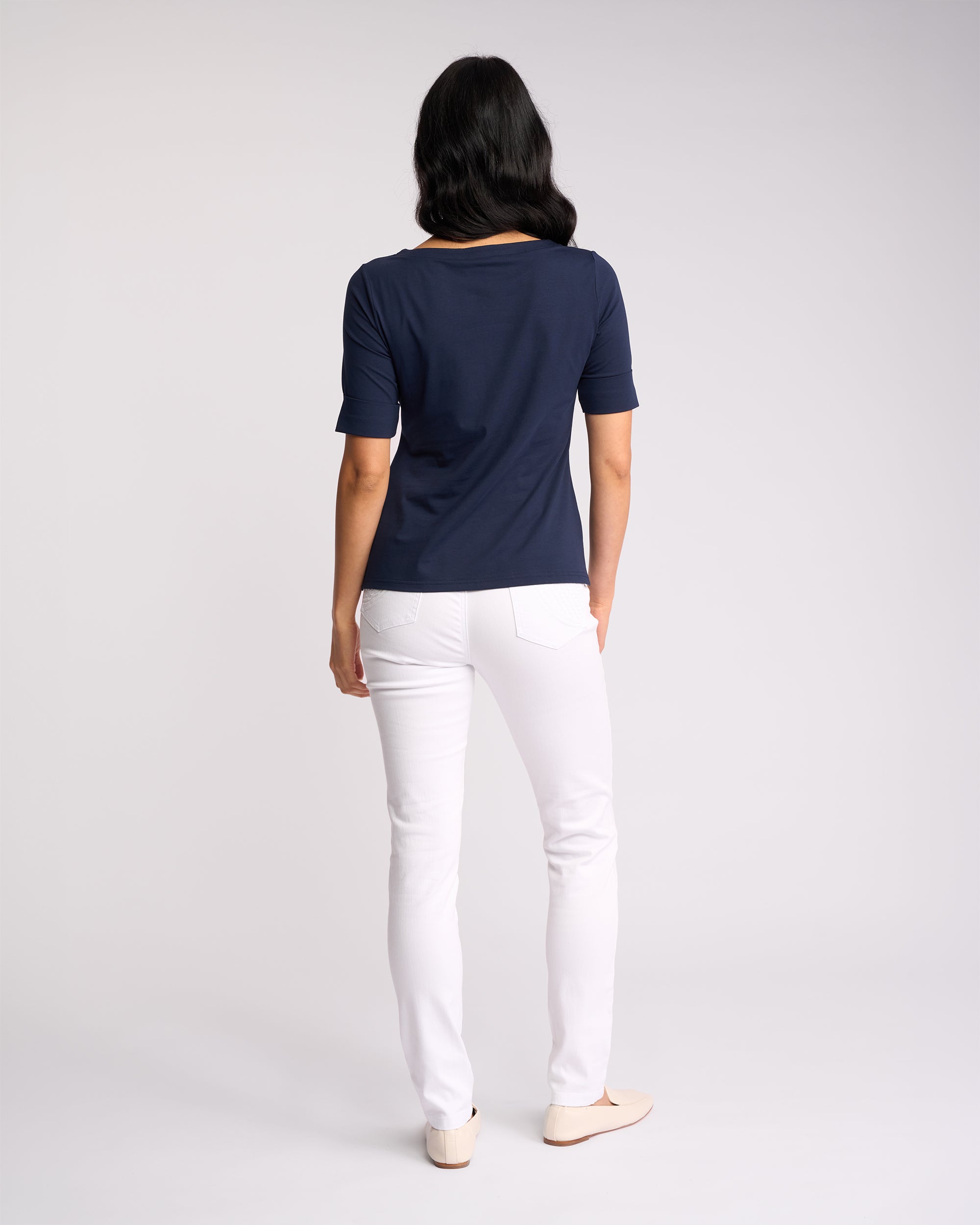 A woman, seen from behind, wears the Vassalli Arden Top - Navy with white pants and light shoes against a plain light background.