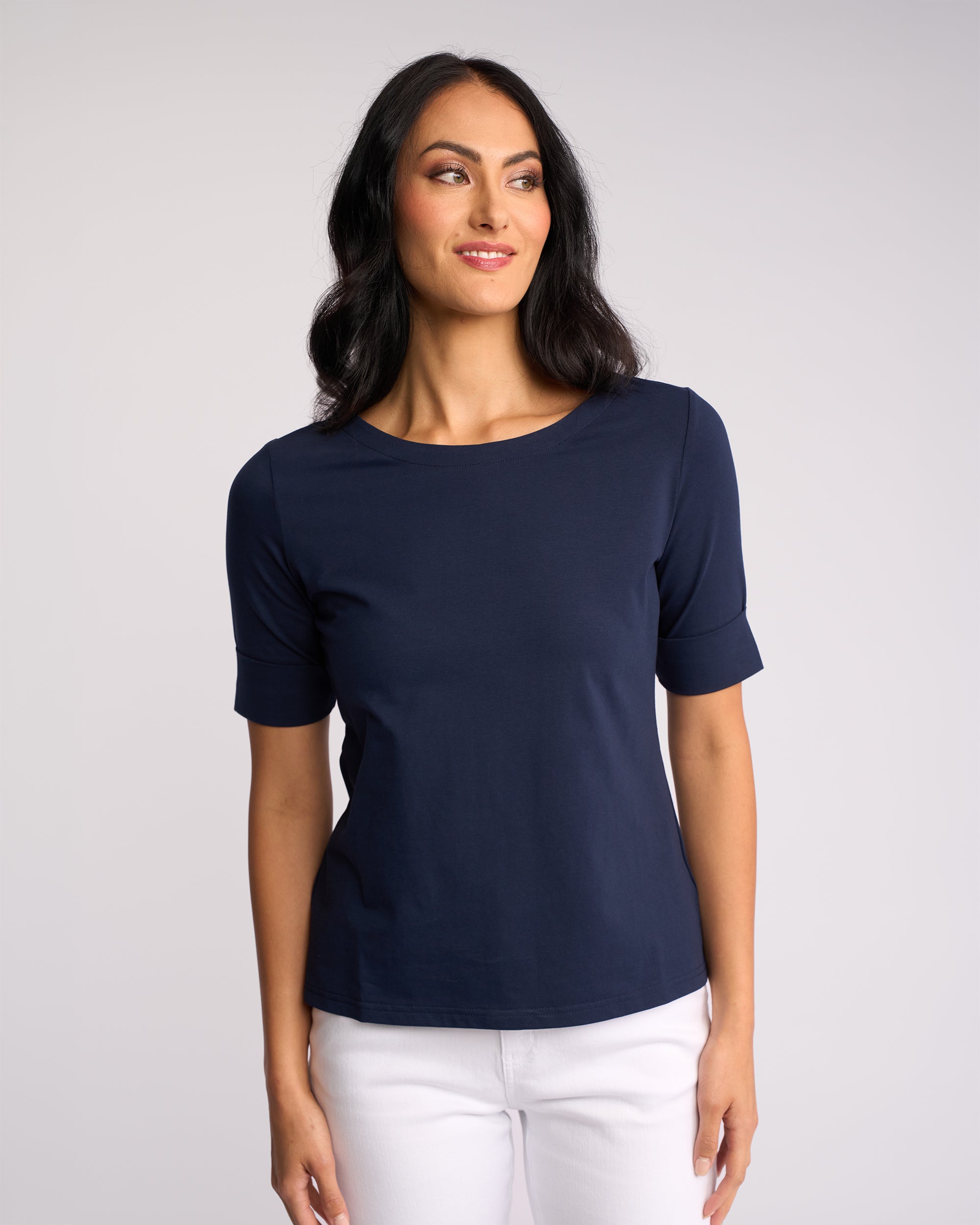 A woman with long dark hair wears the Vassalli Arden Top in navy, made from stretch fabric, paired with white pants. She stands against a plain light background, smiling slightly and looking to her left.