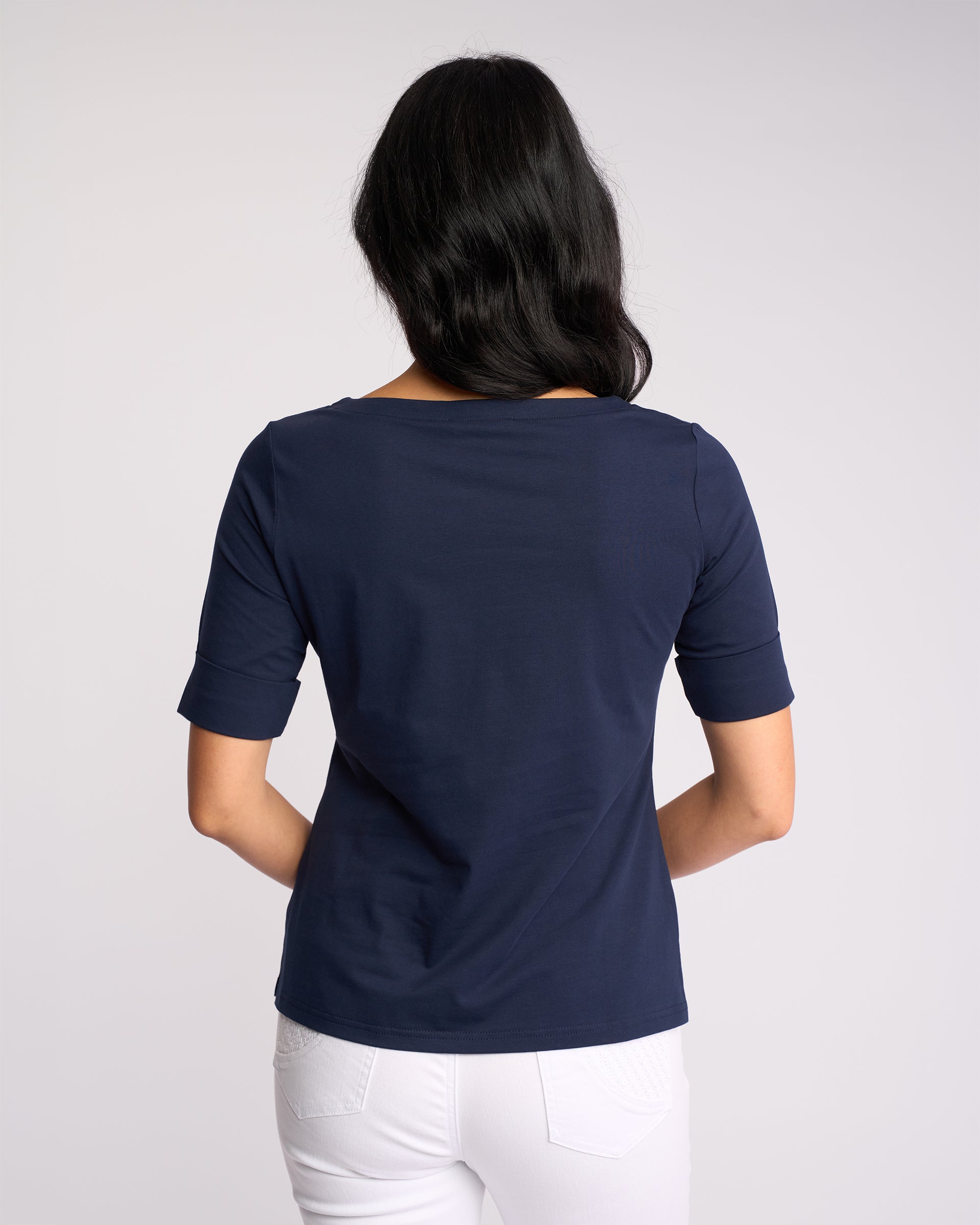 A woman with long dark hair faces away from the camera, wearing the Vassalli Arden Top - Navy and white pants against a plain light background.