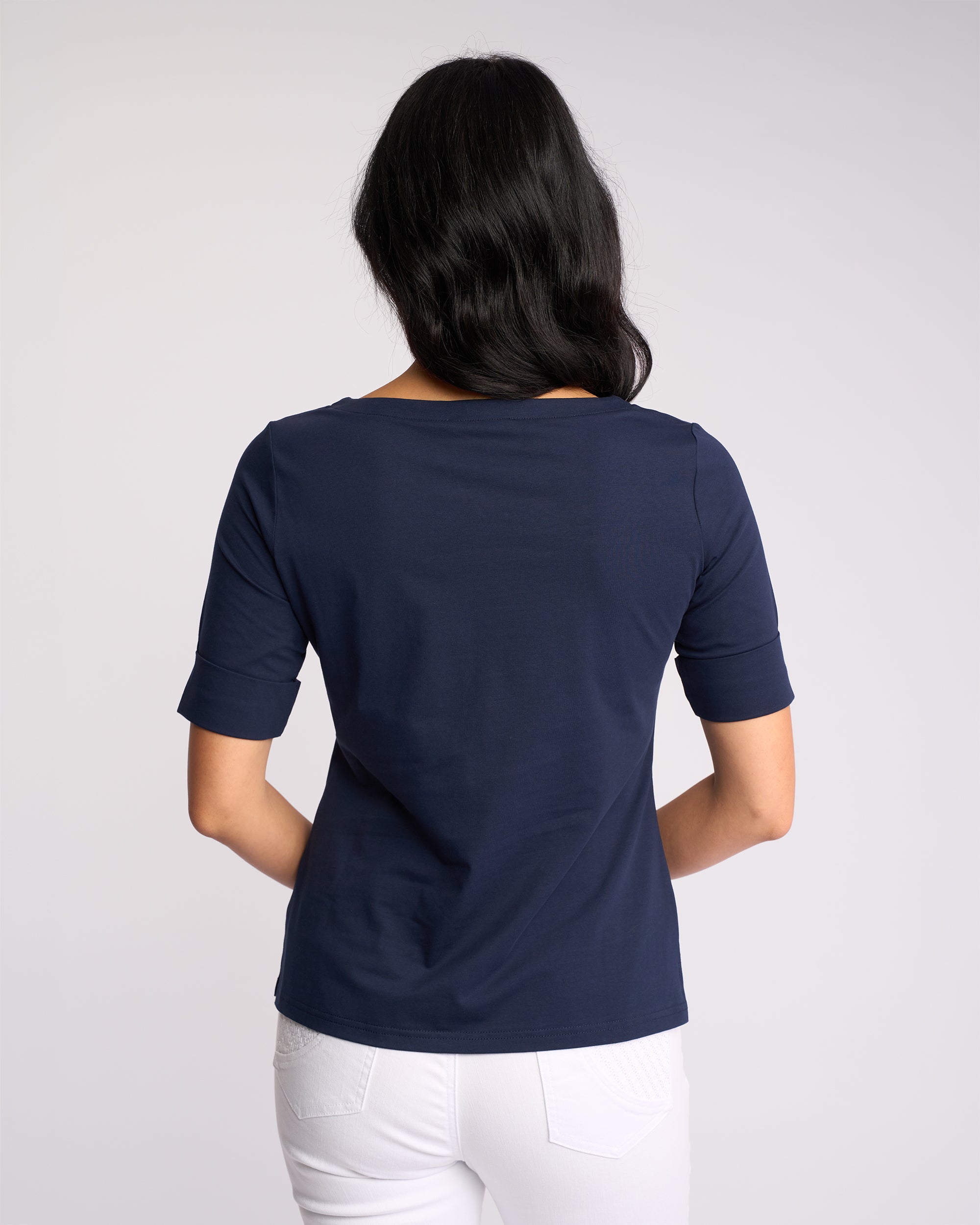 A woman with long dark hair faces away from the camera, wearing the Vassalli Arden Top - Navy and white pants against a plain light background.