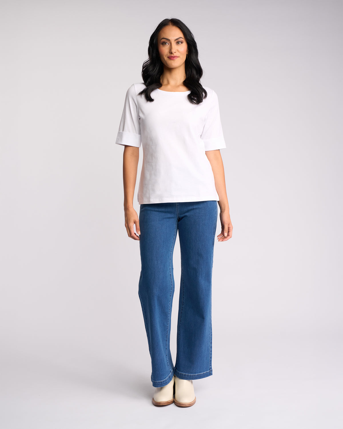 A woman with long dark hair faces forward, wearing the Vassalli Arden Top—plain white with a boat neckline—paired with blue jeans and light shoes, set against a light gray background.